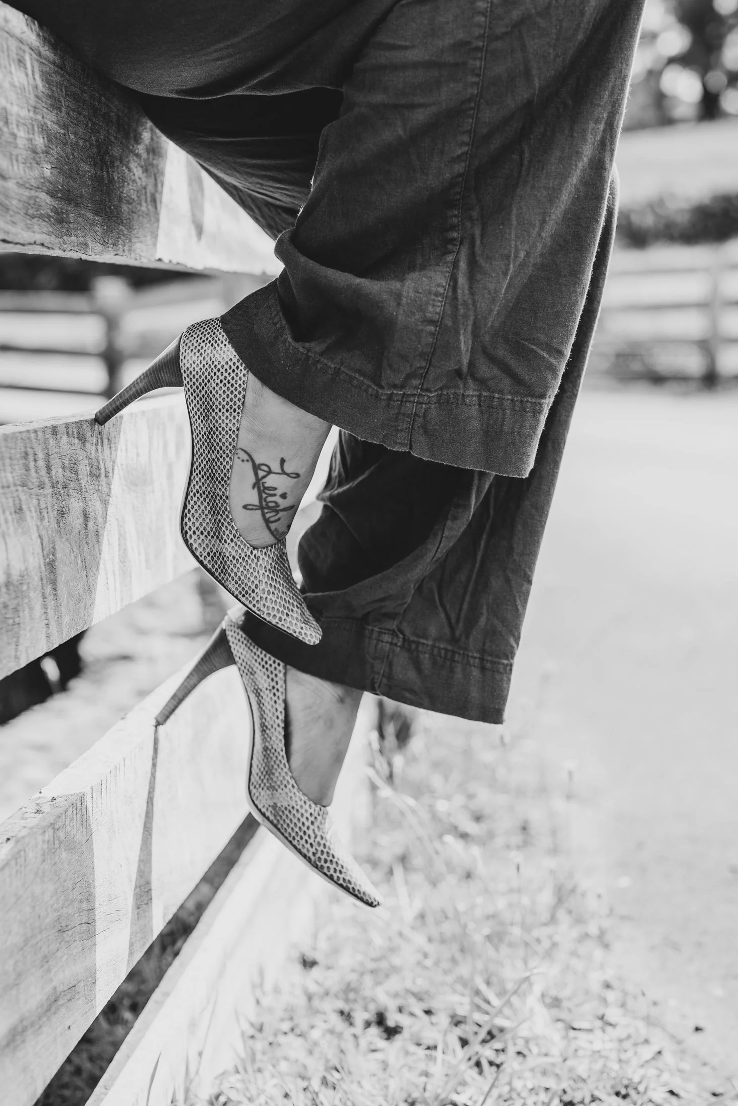 Close-up of a person wearing high-heeled shoes with a tattoo on their ankle, hanging from a wooden fence, in black and white for her maui editorial photographer.