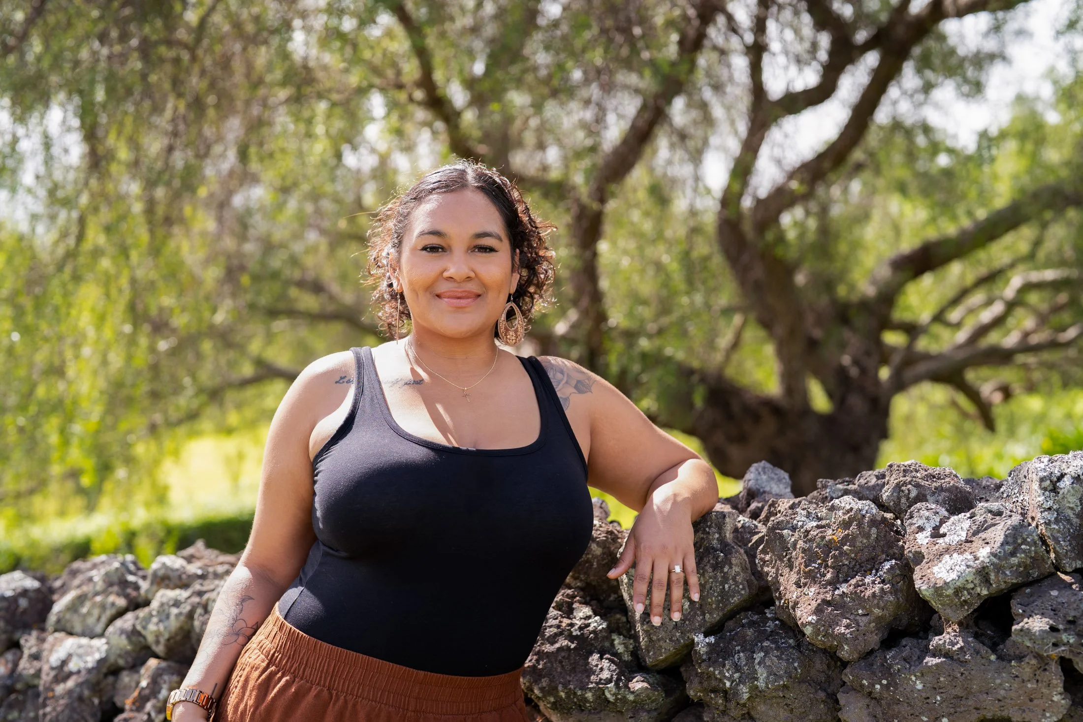 A woman standing outdoors near a stone wall, smiling, with a large leafy tree in the background during daytime during her maui personal branding photographer session.