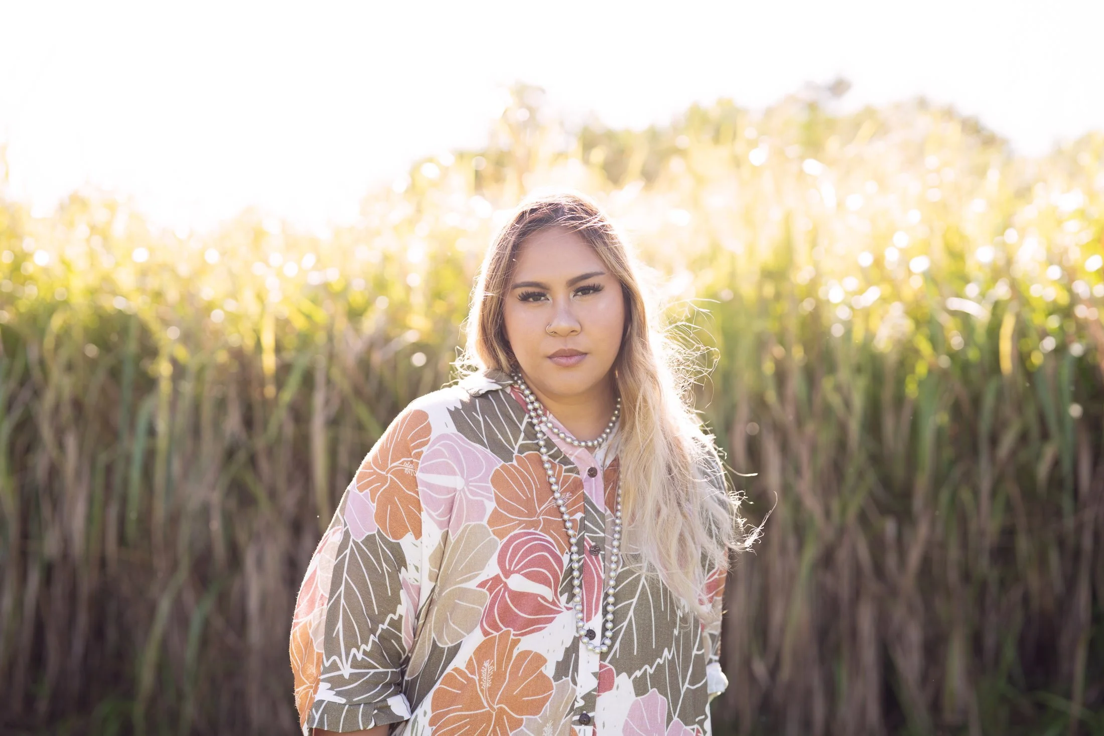 A woman with blonde hair and a floral-patterned blouse standing outdoors in a field of tall grass or crops, illuminated by sunlight from behind during her personal brand photography.