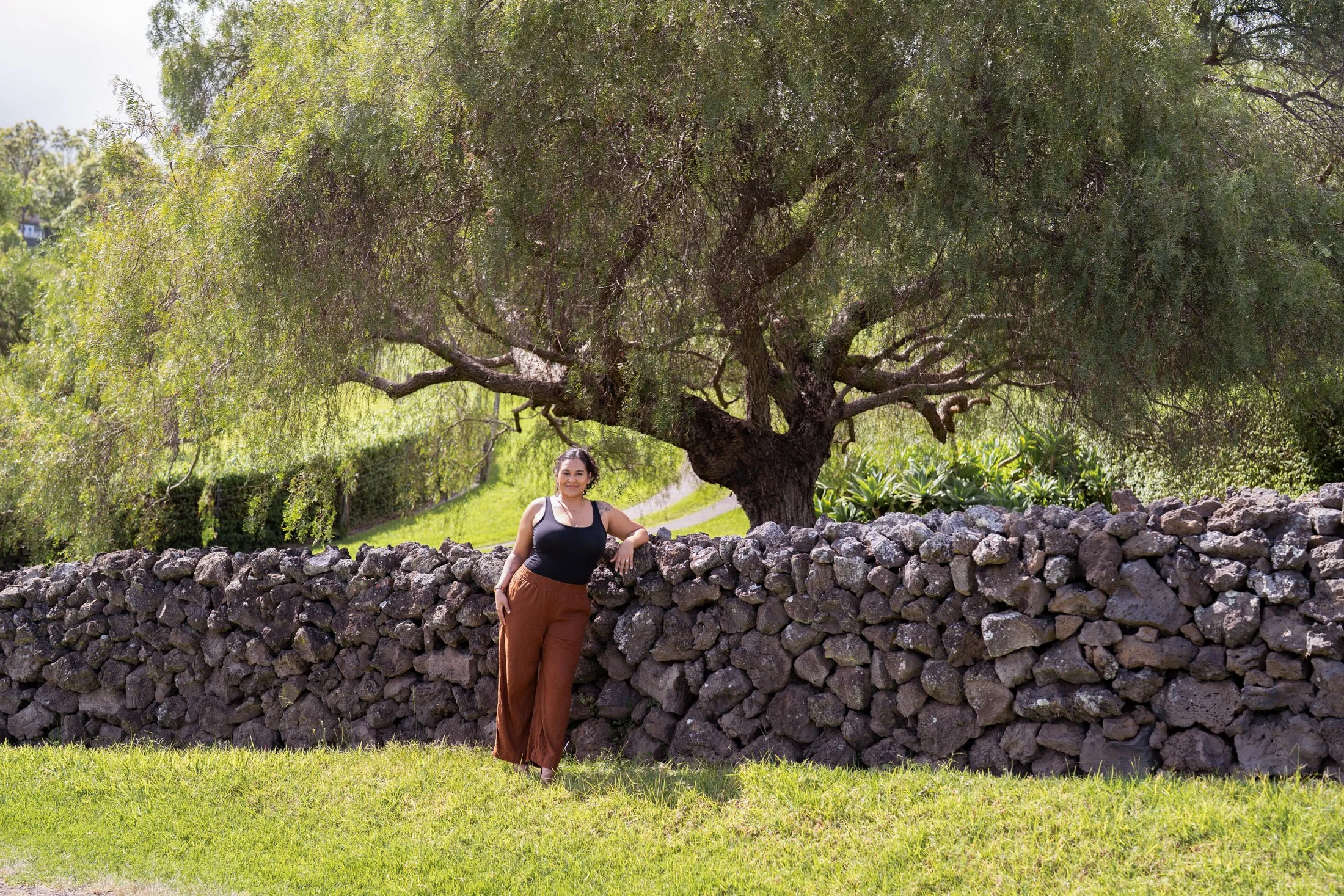 Woman in black top and brown pants leaning against a stone wall under a large, leafy tree in a lush, green outdoor setting during her branding photoshoot maui.