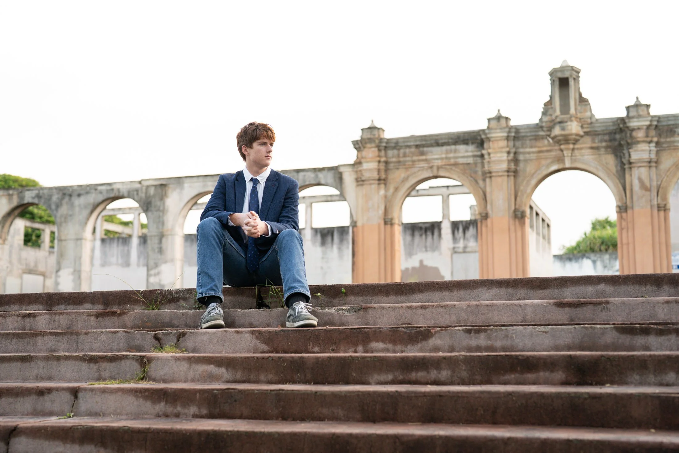 A young man in a business suit sitting on stone steps outdoors with an archway structure in the background of his entrepreneur photoshoot.