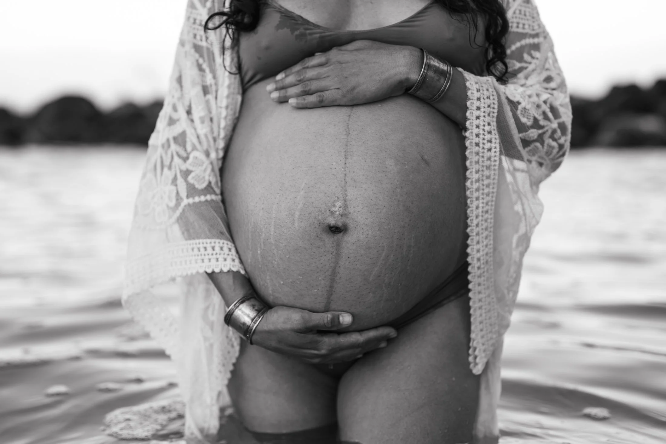 A pregnant woman in a lace cover-up, standing in water at the beach, holding her belly with both hands for her intimate portrait photography.