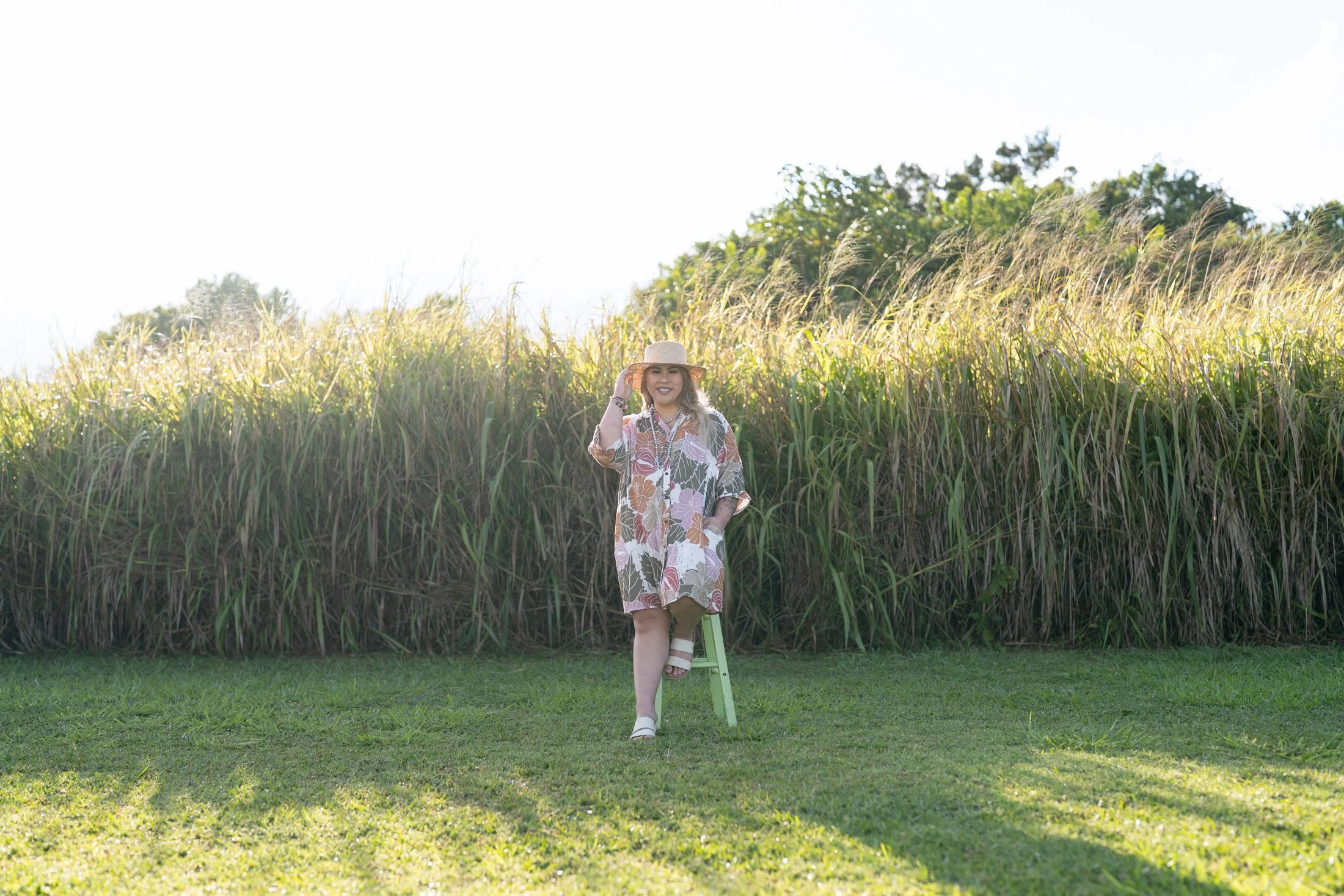 A woman in a floral dress and wide-brimmed hat standing on a stool in front of tall grass in a sunny outdoor setting for her creative business portraits.