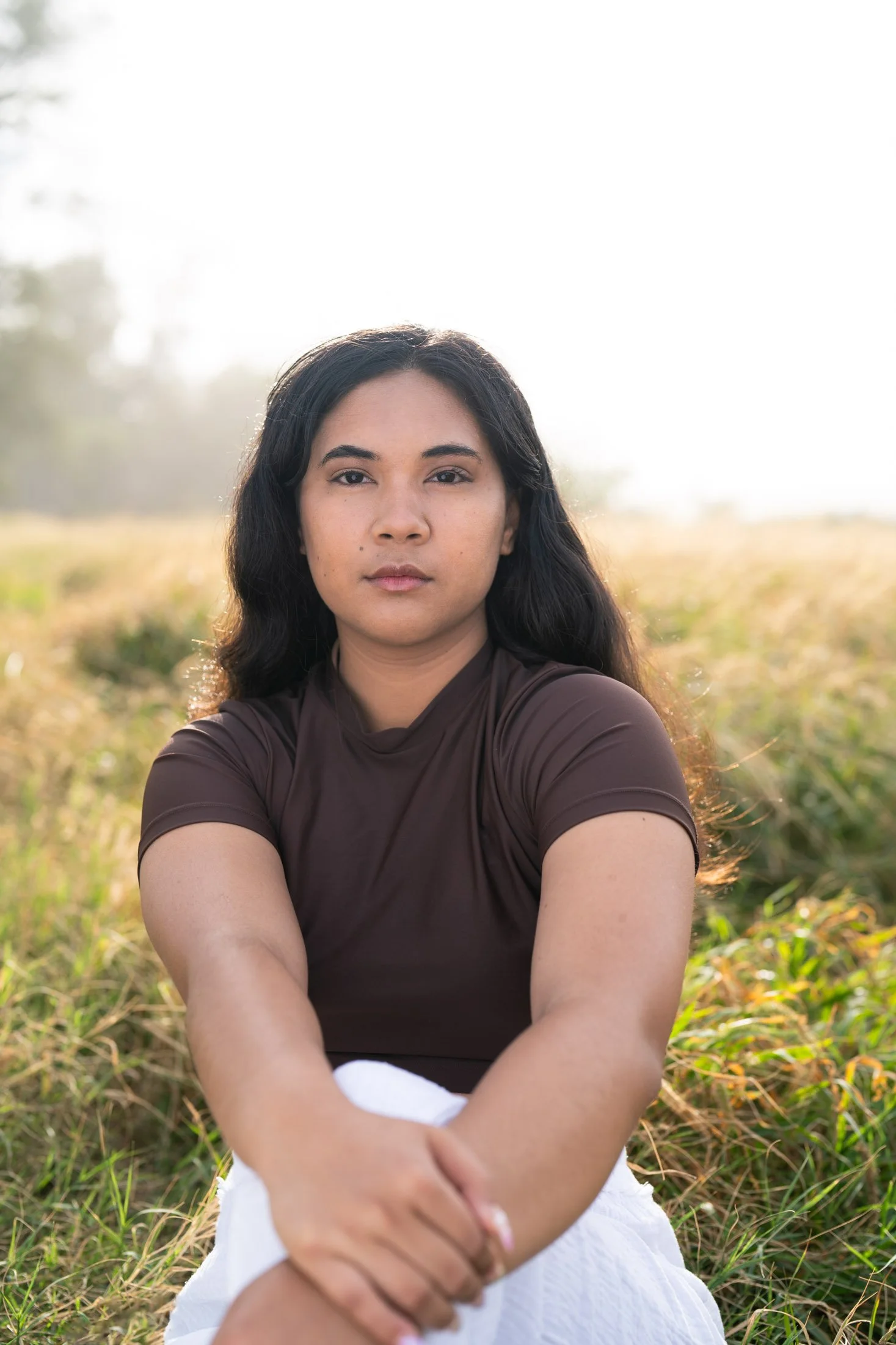 A woman with long black hair sitting in a grassy field during sunset, wearing a brown top and white skirt, with a calm expression for her branding photoshoot maui.
