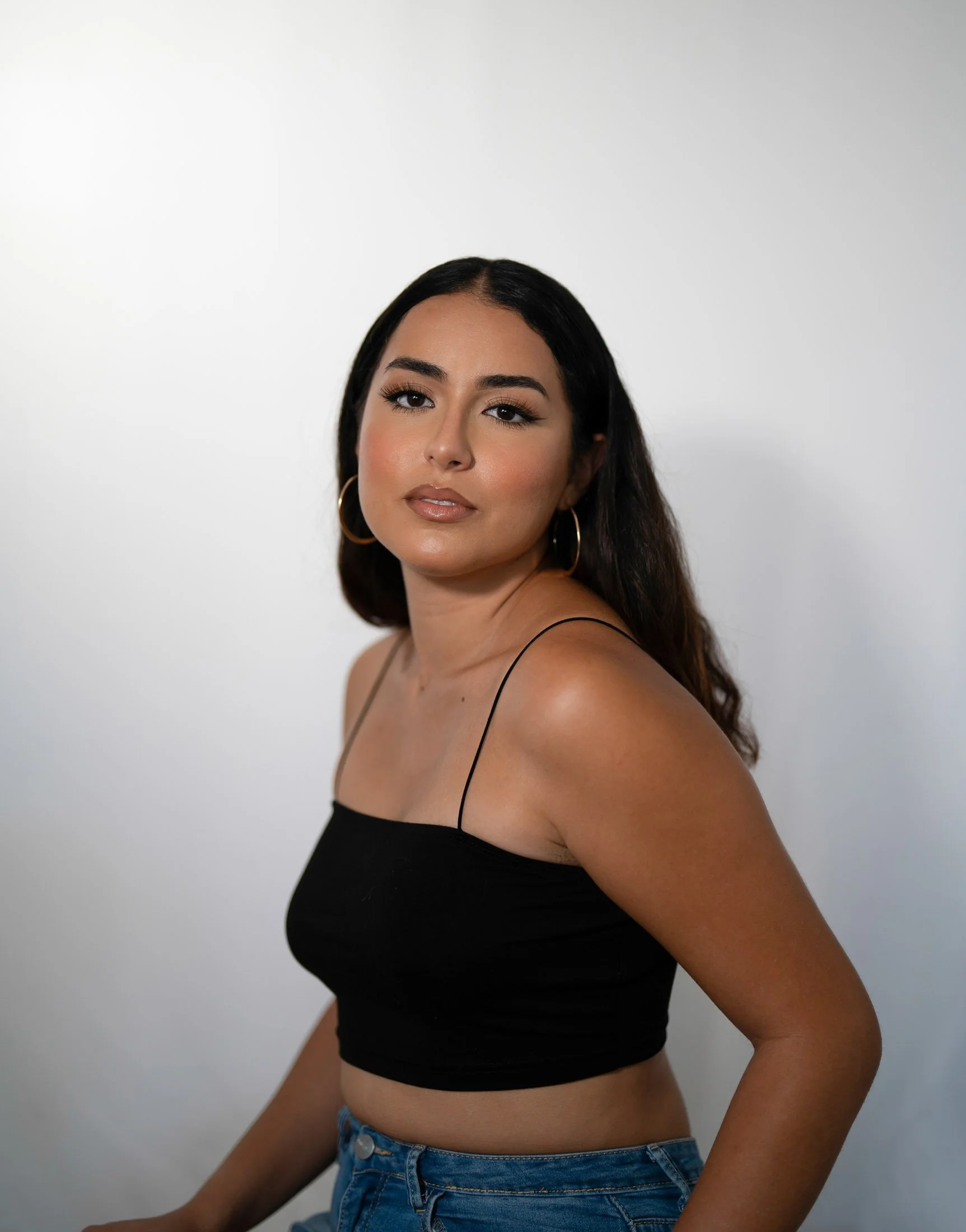 A young woman with long dark hair, wearing a black spaghetti strap crop top and blue jeans, poses against a plain white background during her creative business portraits.
