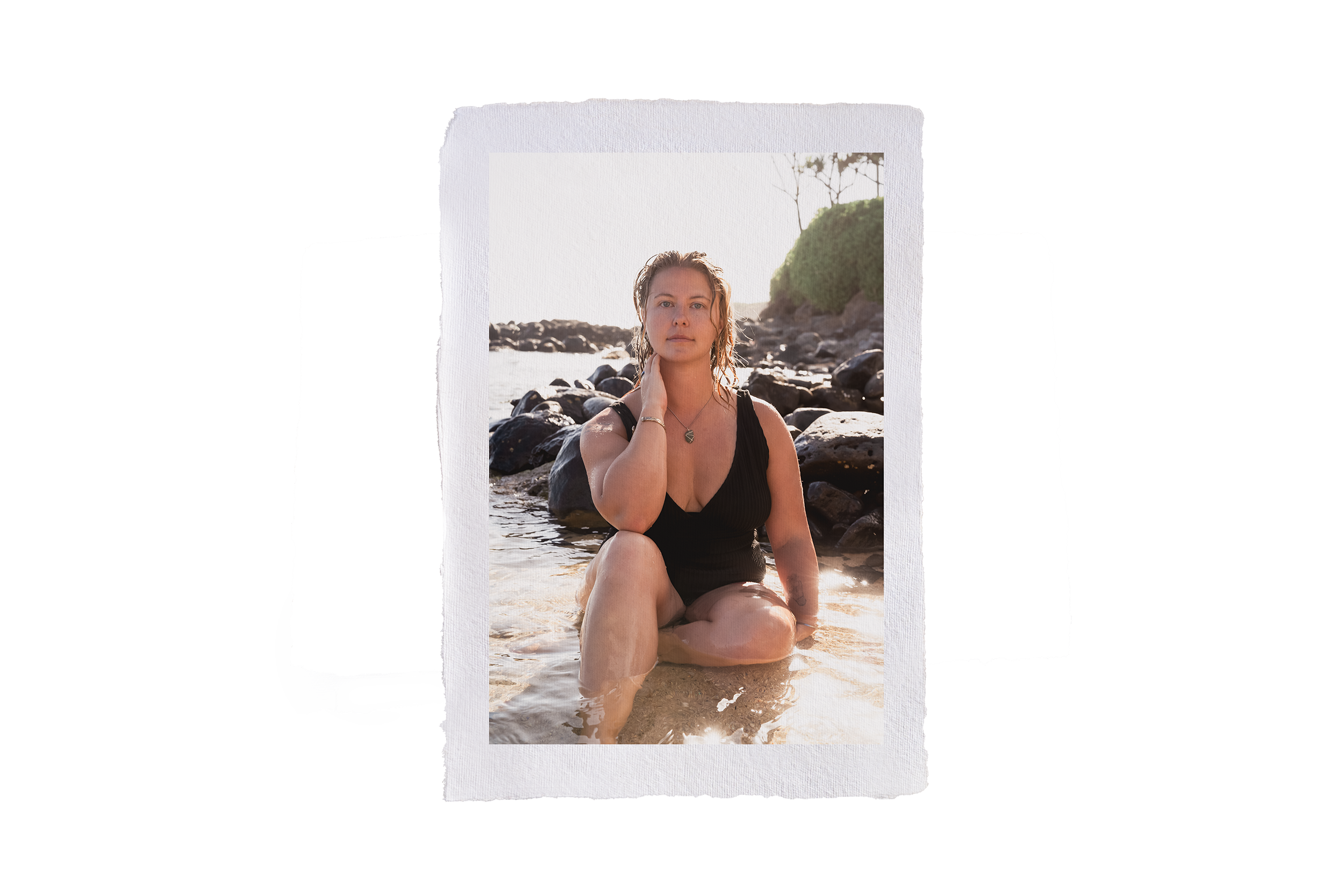 A woman sitting in shallow water on a rocky beach in the morning light, wearing a black swimsuit and jewelry, with water and rocks around her and a green bush in the background during her intimate portrait photography session.