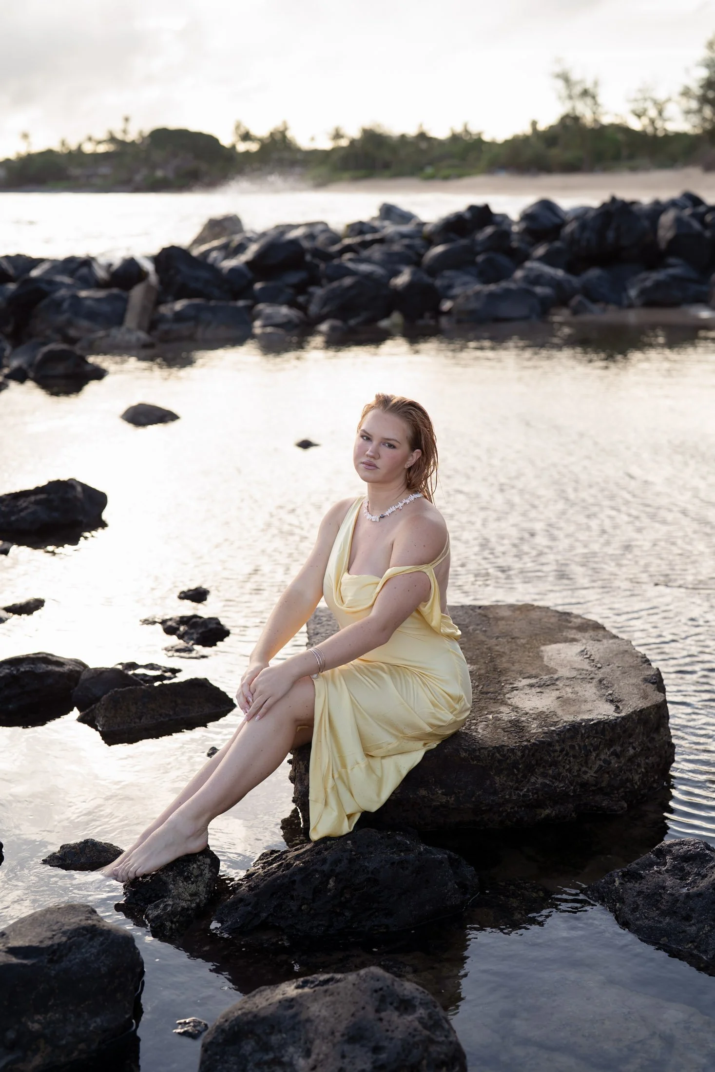 A woman in a yellow dress sitting on a large rock in a cove at sunset for her editorial portrait photography session. 