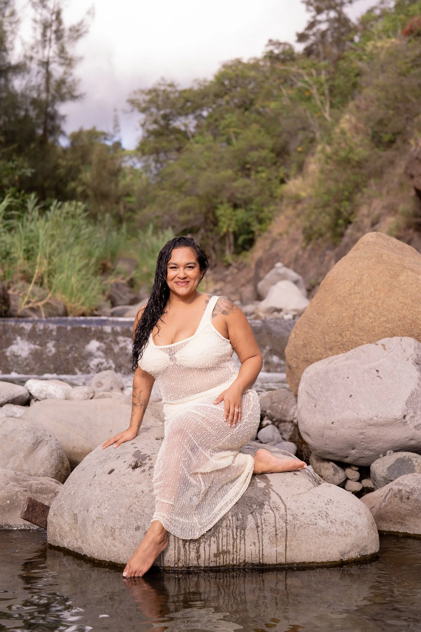Woman in a white dress sitting on a large rock in a shallow creek, smiling, with lush trees and rocks in the background during her outdoor boudoir photography session.