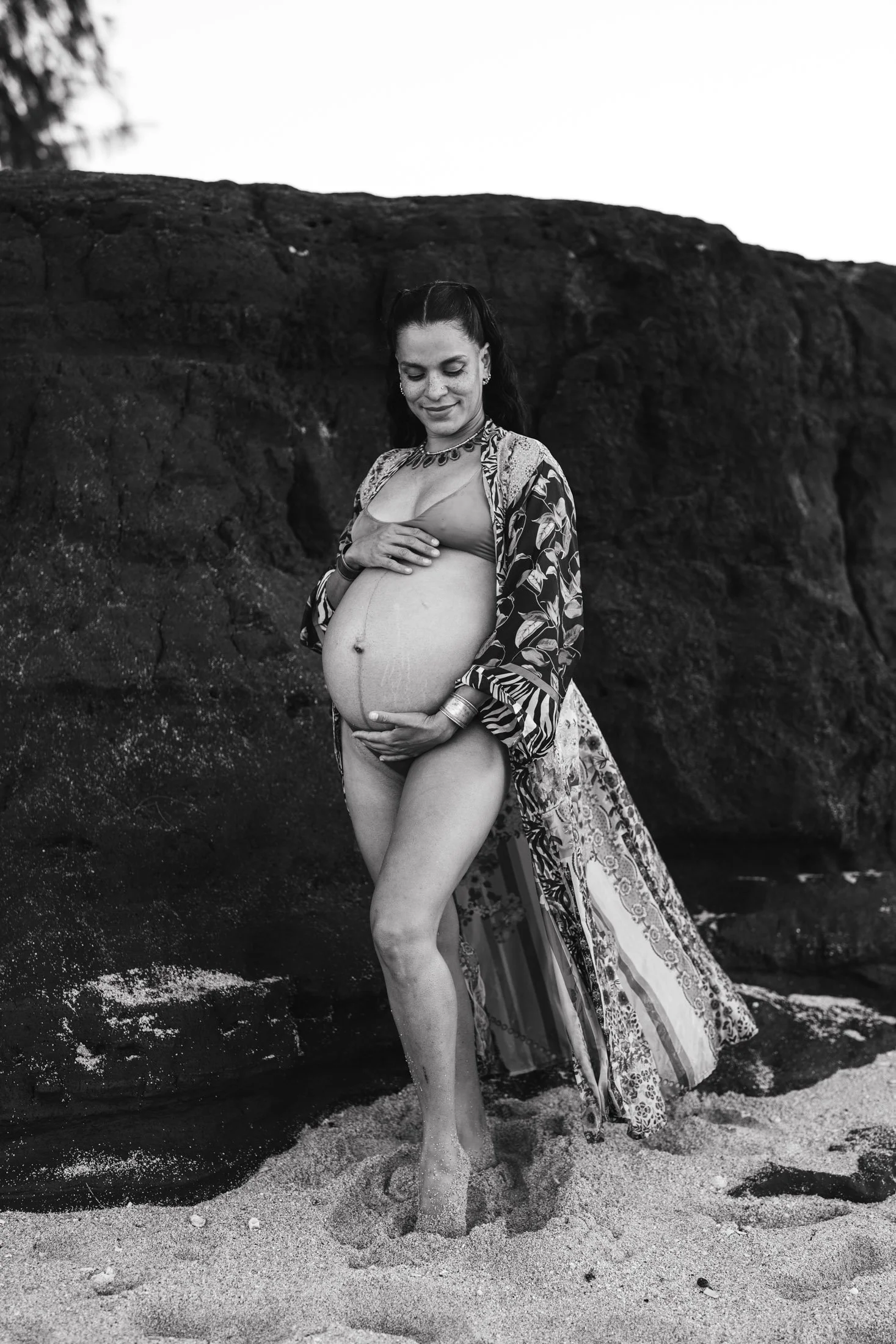 A pregnant woman standing on the beach, touching her belly and smiling, with a large rock formation behind her, in a black and white photo during her natural light portraits maui.
