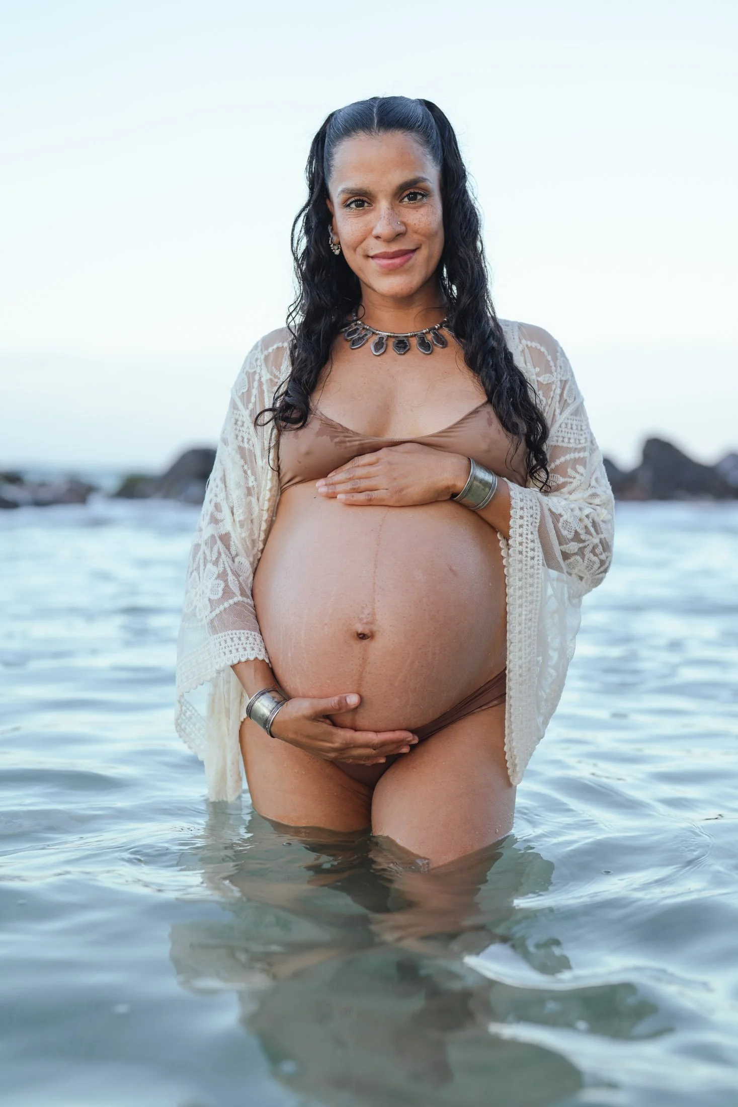 Pregnant woman standing in the ocean, holding her belly with one hand, smiling at the camera, wearing jewelry and a lace cover-up, with rocks and a clear sky in the background during her artistic photoshoot.