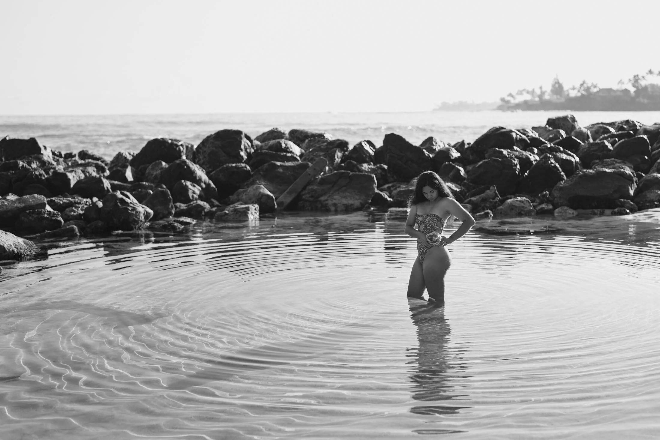 A woman standing in shallow ocean water near rocks, holding a hat, in a black and white photo with a distant shoreline and trees in the background during her outdoor portrait photography session.