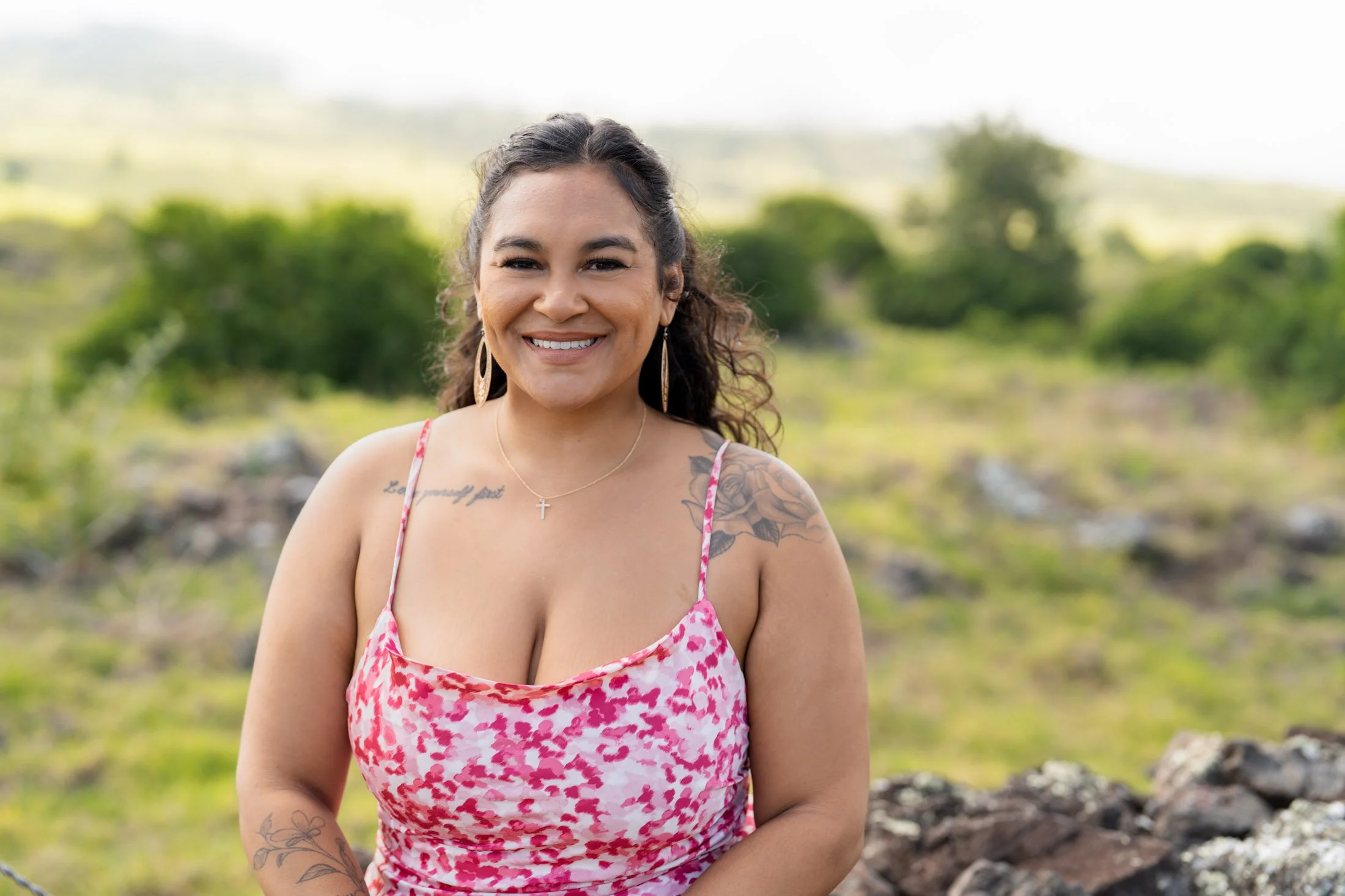 Smiling woman with tattoos, wearing a pink patterned dress and jewelry, standing outdoors in a green landscape during her entrepreneur photoshoot.