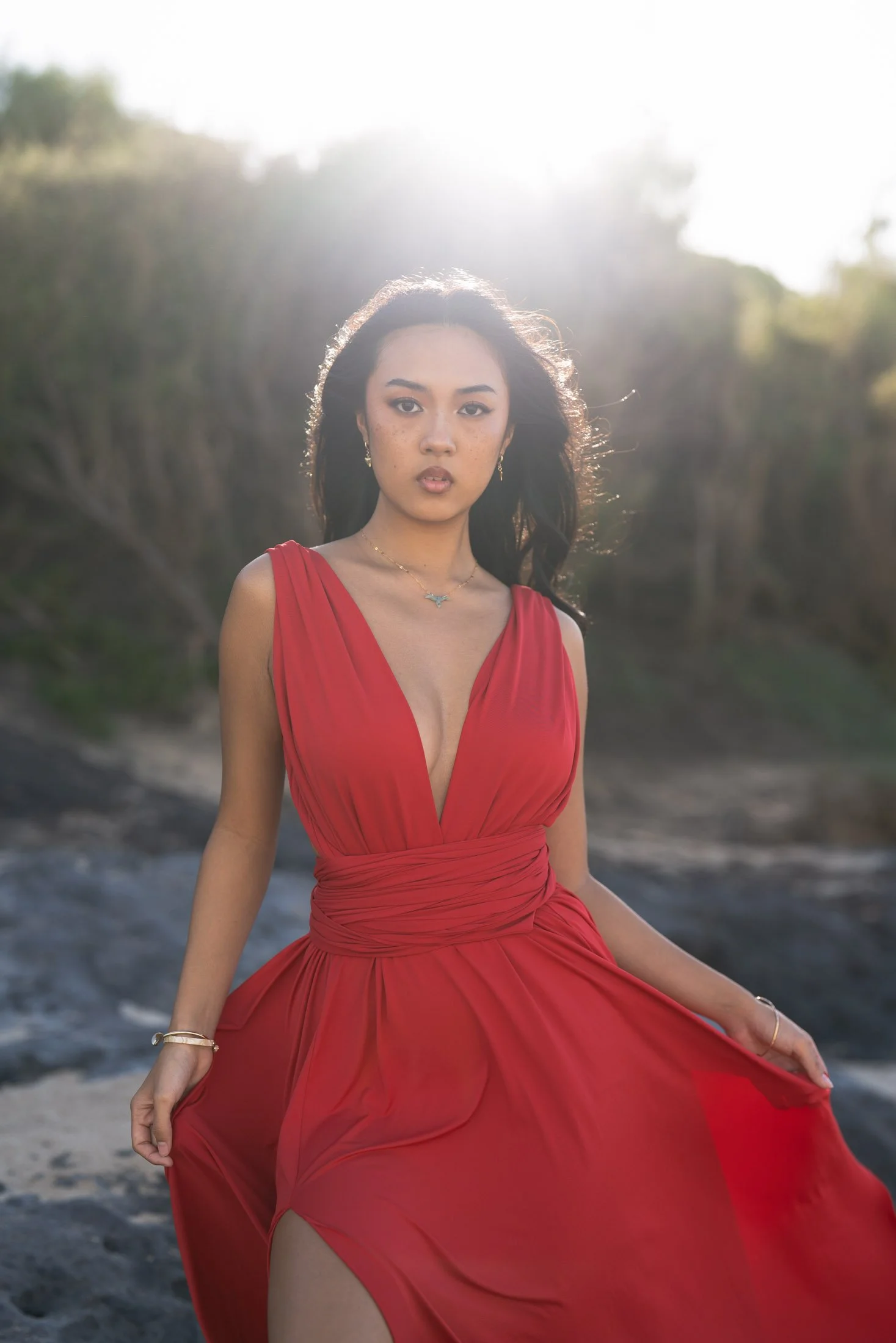A woman in a red gown standing outdoors, backlit sunlight creating a glow around her, with a natural background of trees and rocky terrain for maui editorial photographer.