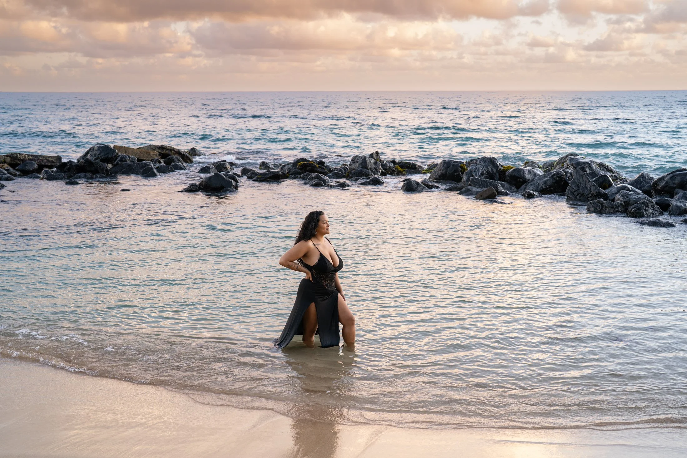 Woman in a black dress standing in shallow ocean water near rocks during sunset during her outdoor boudoir photography session