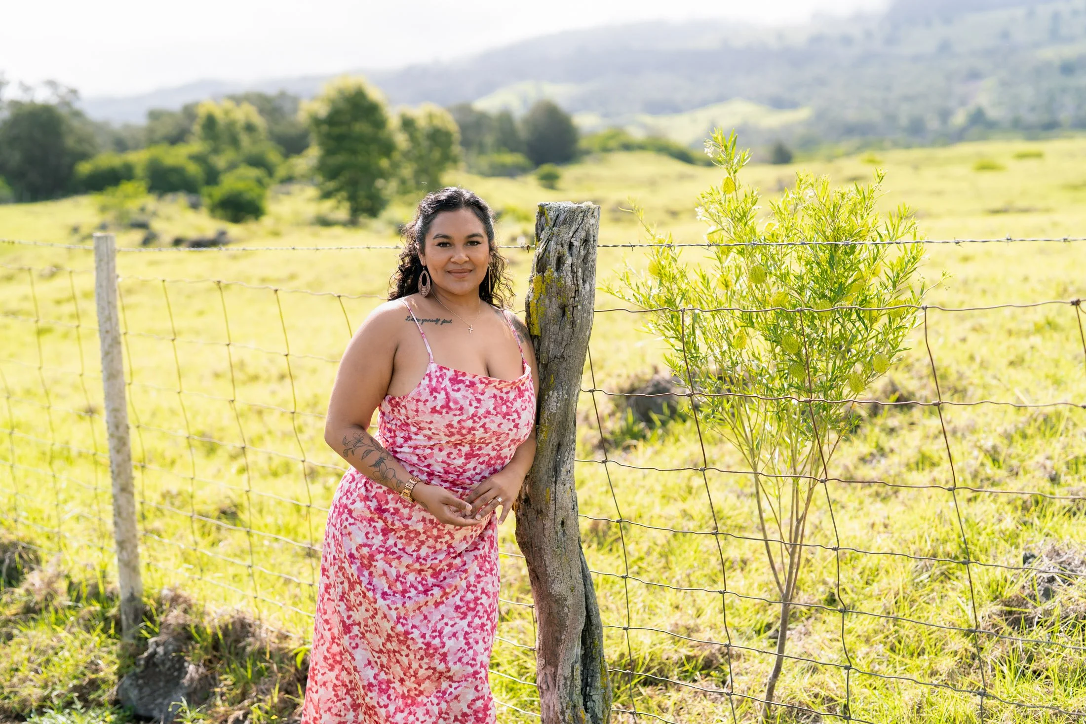 A woman stands outdoors next to a wooden fence post and wire fence, smiling at the camera. The background features a grassy field, green trees, and rolling hills under a partly cloudy sky for her entrepreneur photoshoot.