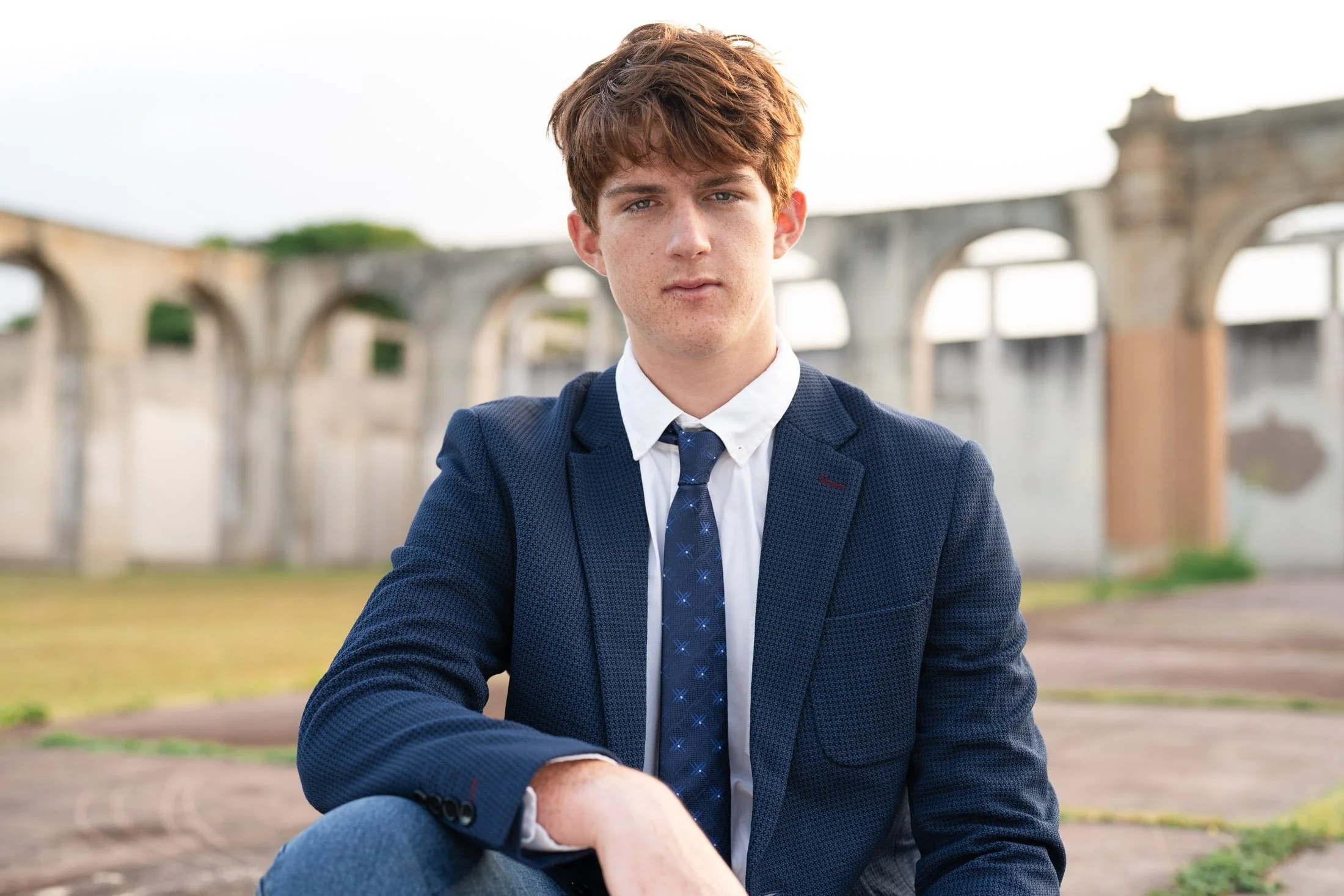 A young man with reddish-brown hair wearing a navy blue blazer, white shirt, navy blue tie, and gray pants, sitting outdoors for his maui personal branding photographer.