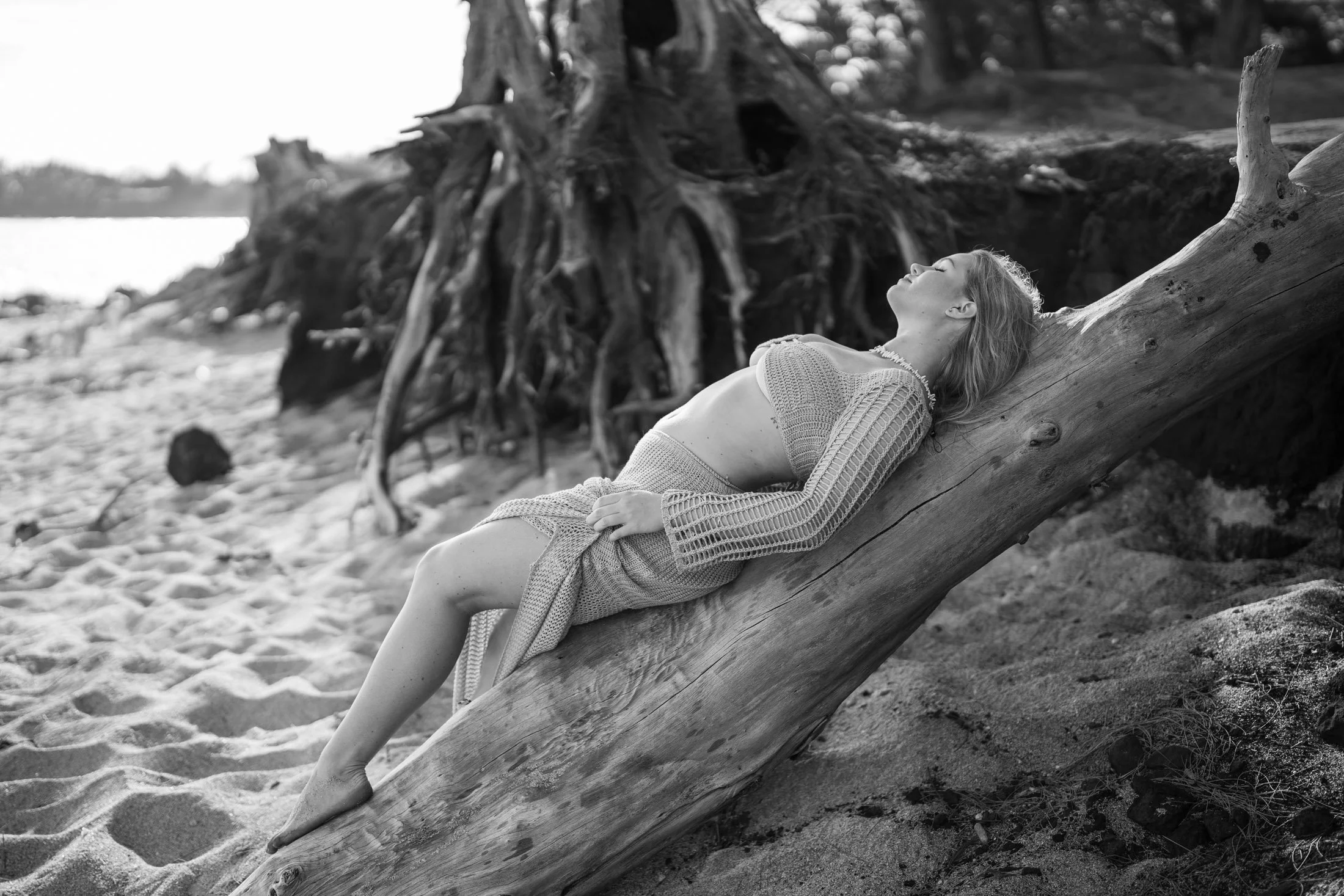 A woman with light-colored hair lying on a slanted log on a sandy beach, wearing a crocheted top and skirt, with her eyes closed and head tilted back during her sensual photoshoot maui.