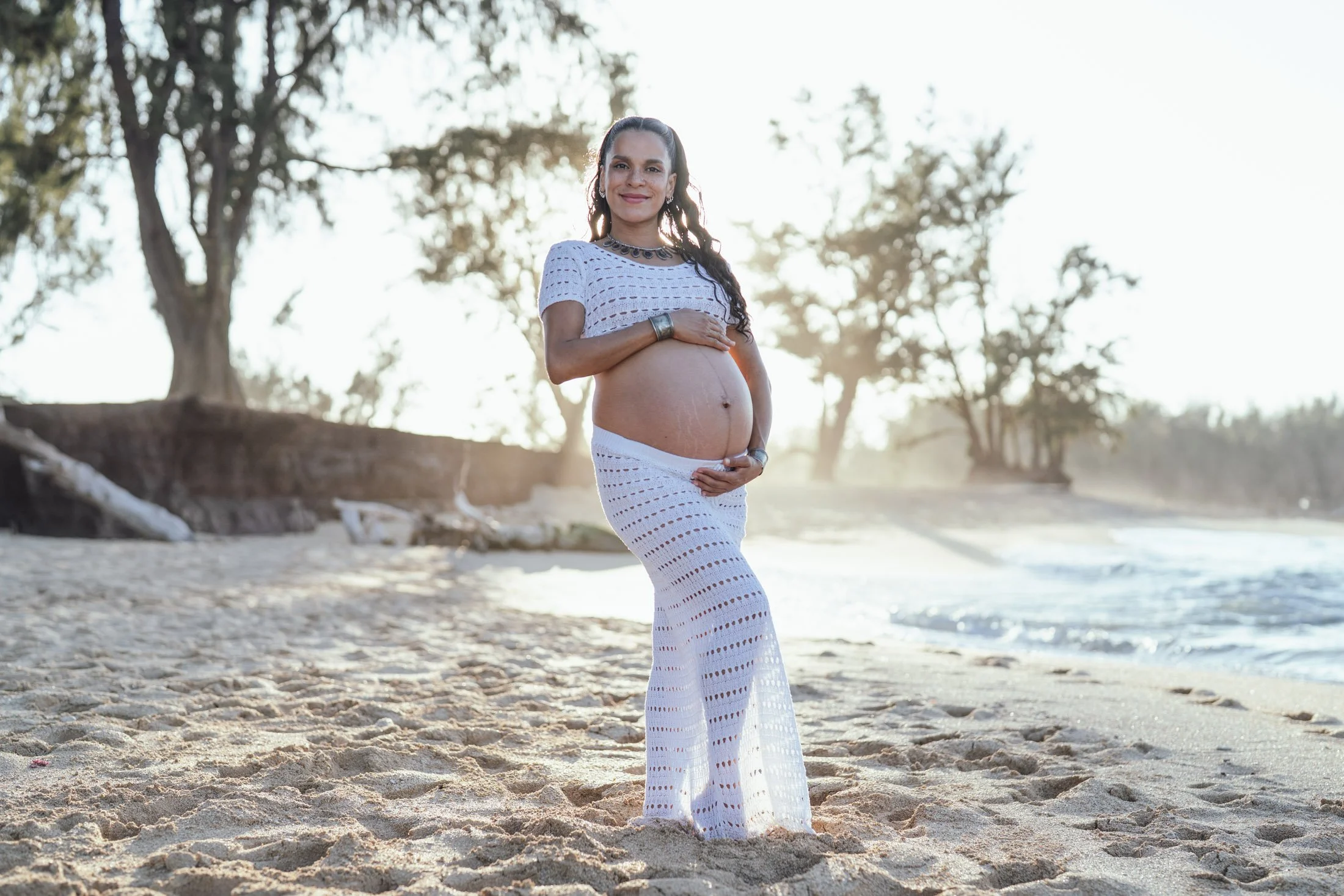 A pregnant woman standing on a beach during sunset, wearing a white crochet dress and jewelry, smiling while holding her belly for her natural light portraits maui.