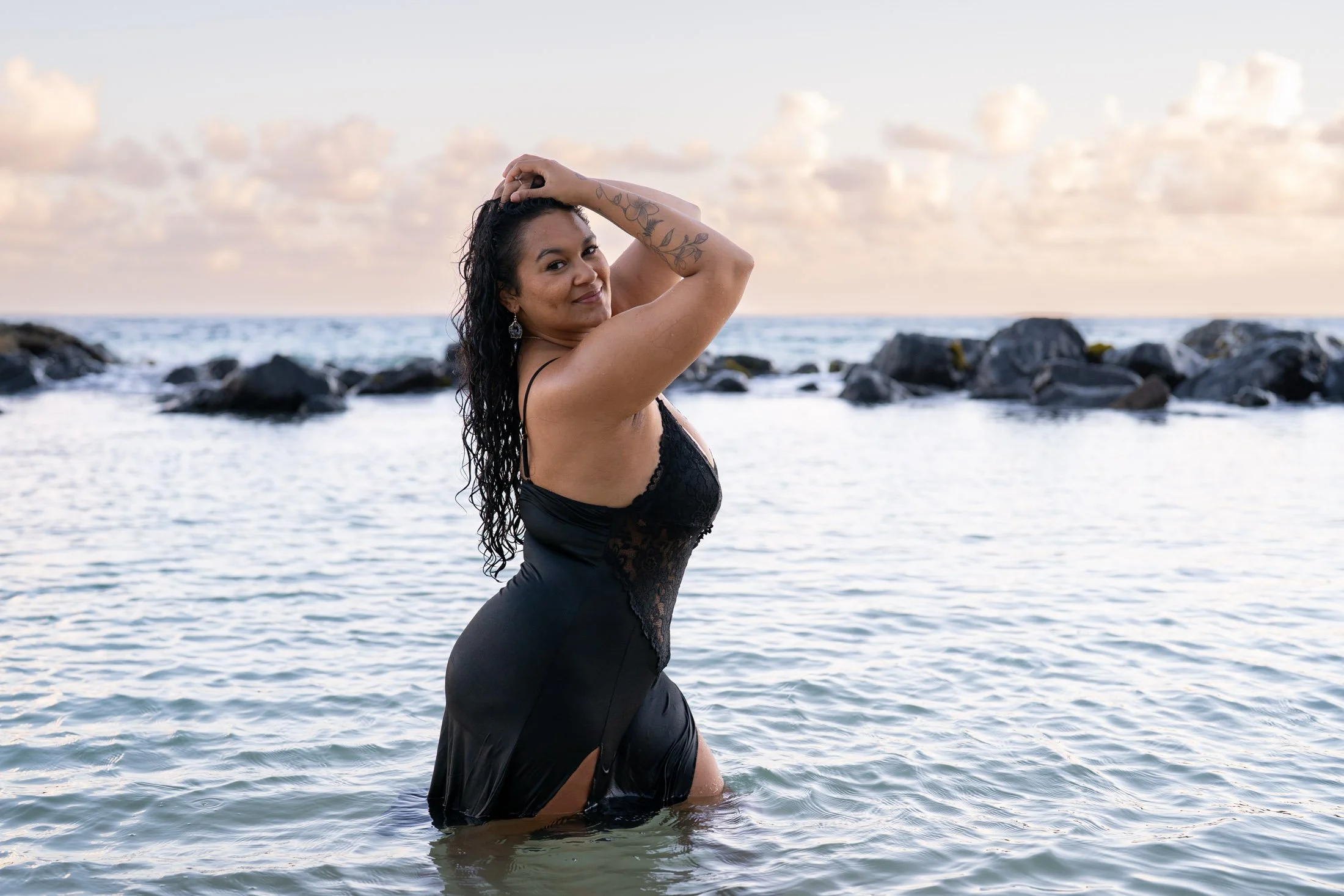 A woman in a black dress standing in the ocean near rocks at sunset, with long wet hair and a tattooed arm, smiling and looking at the camera during her empowerment boudoir session.