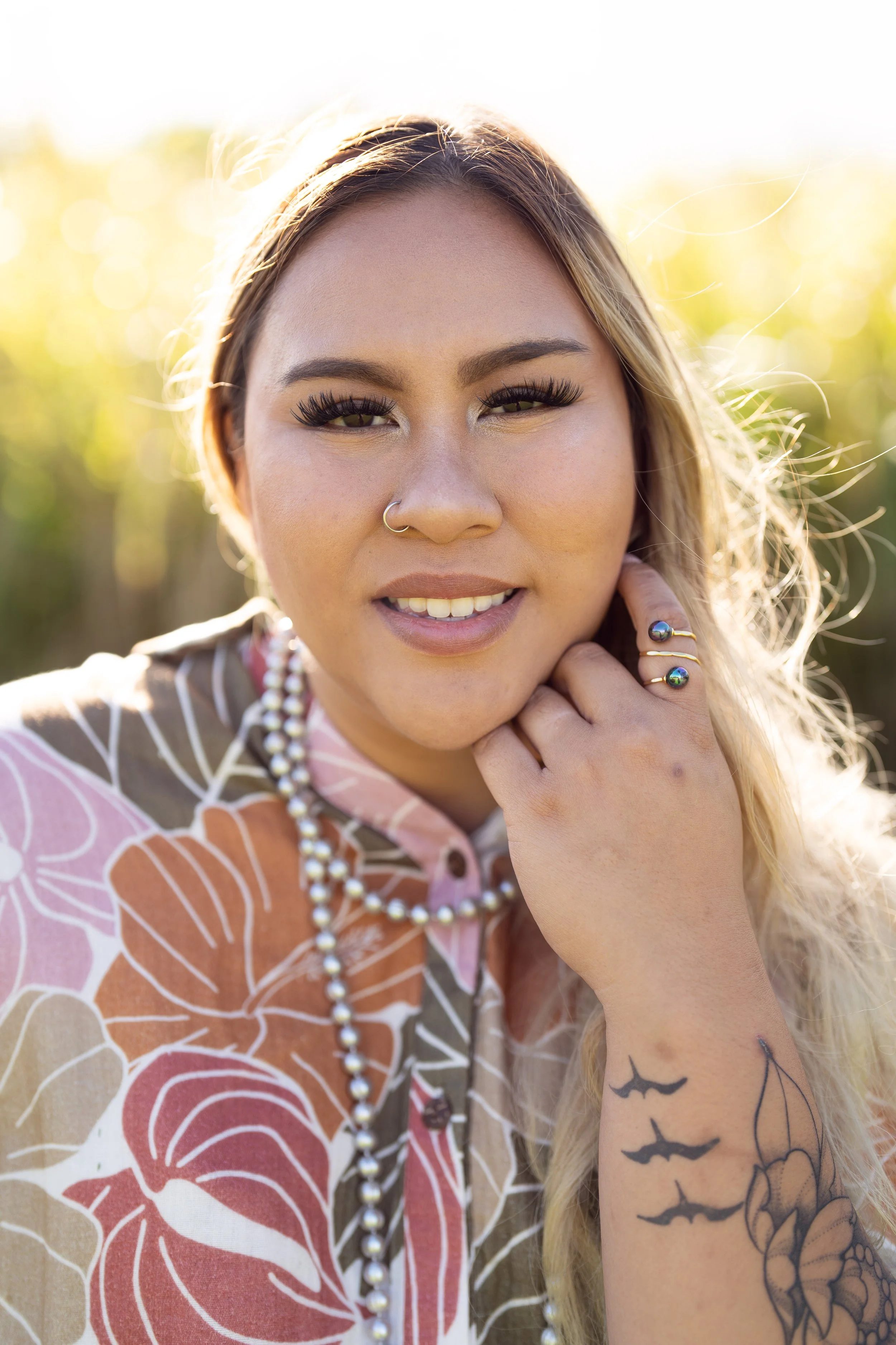A woman with long blond hair, wearing makeup and jewelry, smiling outdoors with a blurred background of greenery and sunlight for her creative business portraits.