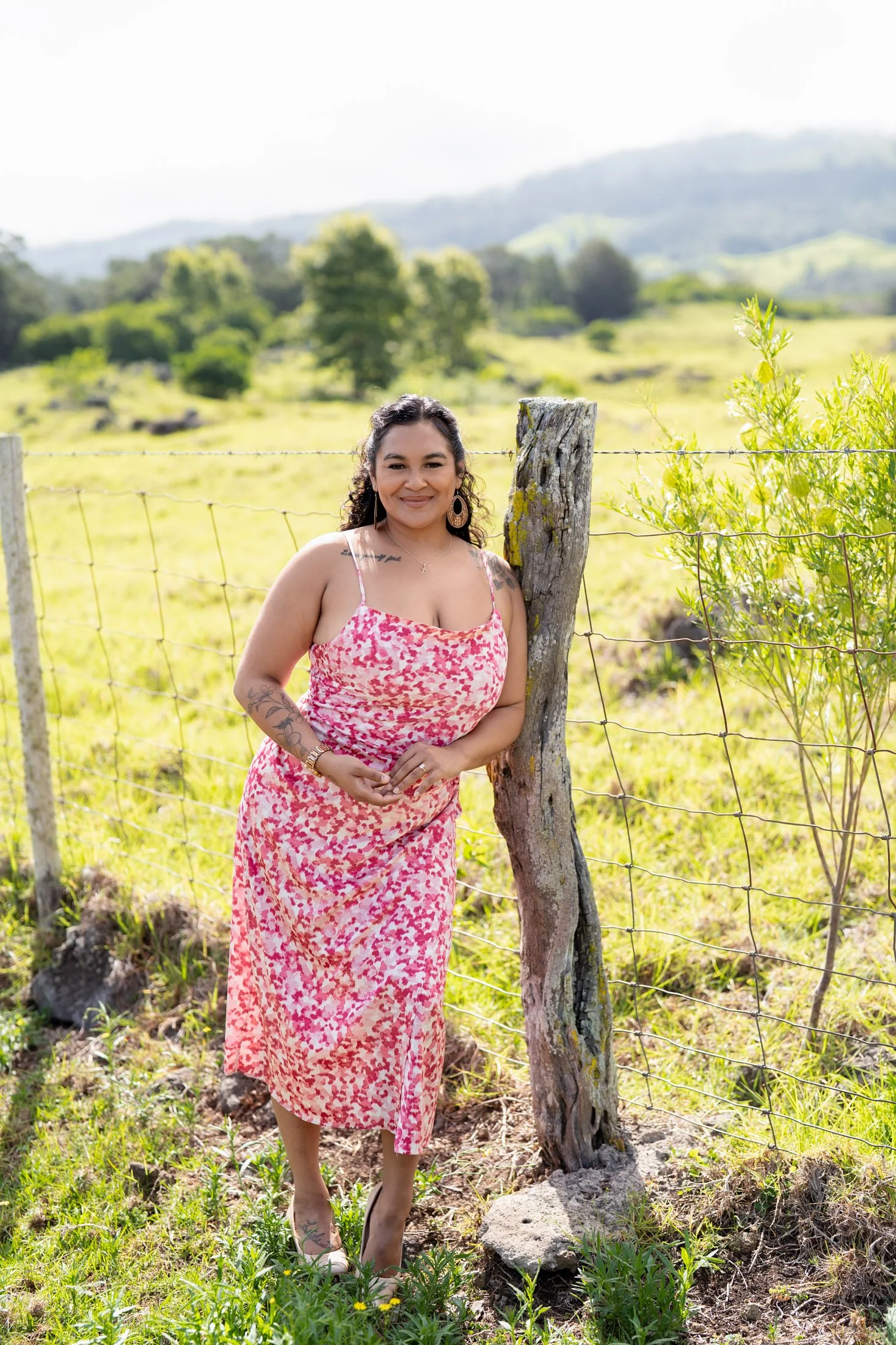 Woman standing outdoors in a pink patterned dress, leaning against a wooden fence post, with green fields and hills in the background during her branding photoshoot maui.