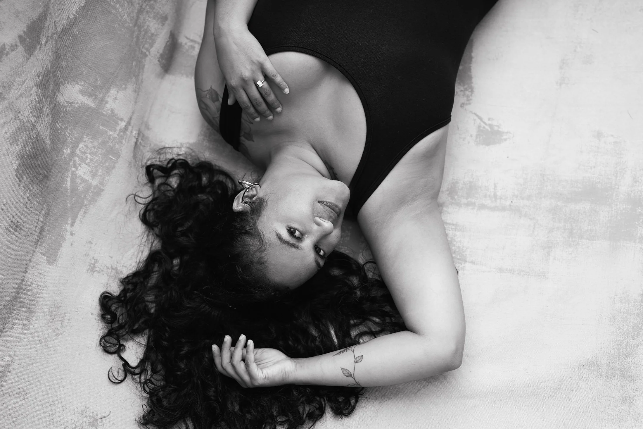 A woman with curly hair lying on her back on a textured surface, wearing a black sleeveless top, with her left hand resting on her chest and her right hand in her hair, looking at the camera during her artistic photoshoot maui.