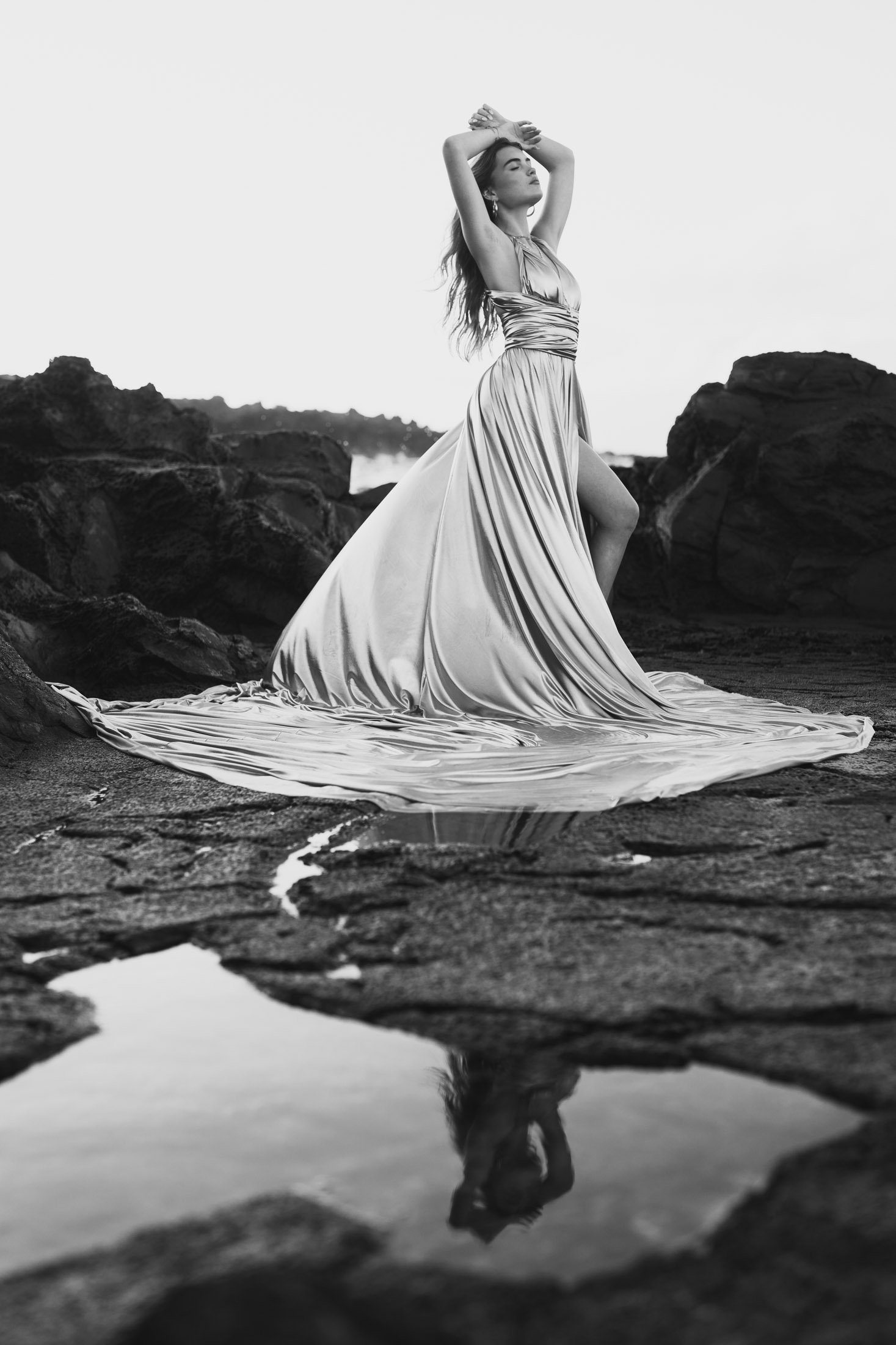 A woman in a flowing gown stands on rocky terrain near water, with her eyes closed and arms raised above her head, her reflection visible in a small pool of water during her artistic photoshoot maui.