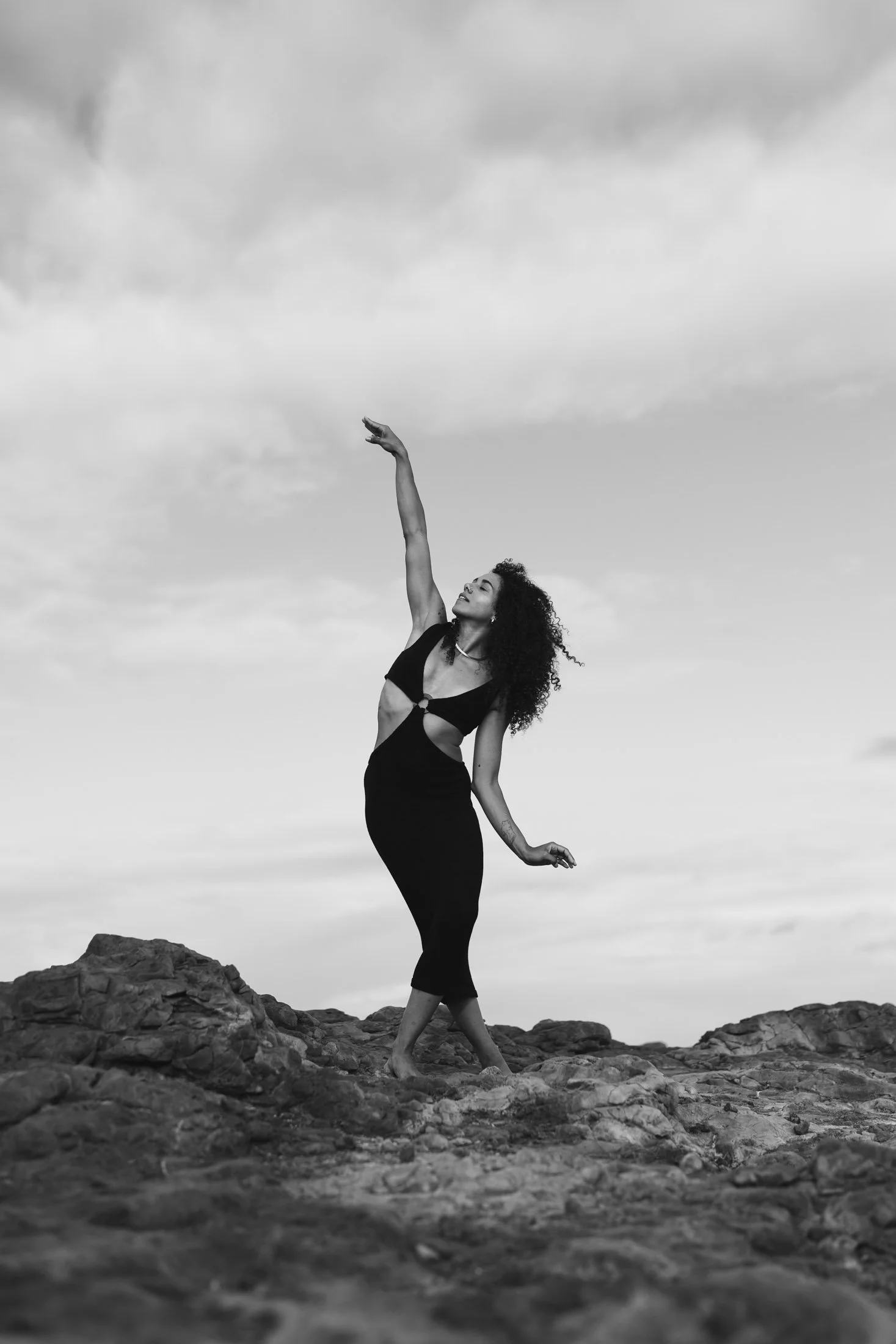 A woman with curly hair dancing barefoot on rocks outdoors, wearing a black dress with cutouts, against a cloudy sky.
