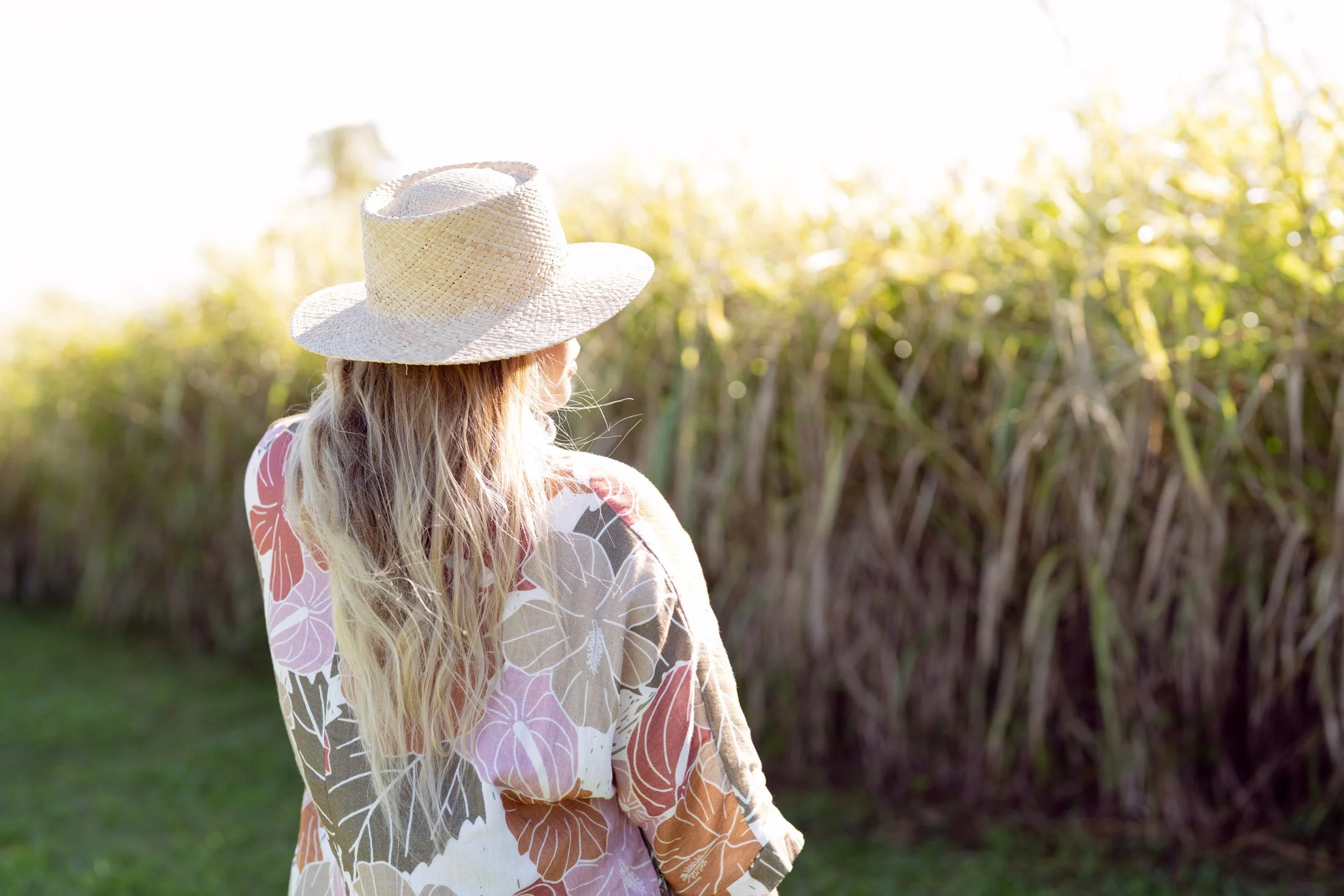 A woman with long blonde hair, wearing a straw hat and a floral blouse, standing outdoors near tall grass for her branding photoshoot maui.