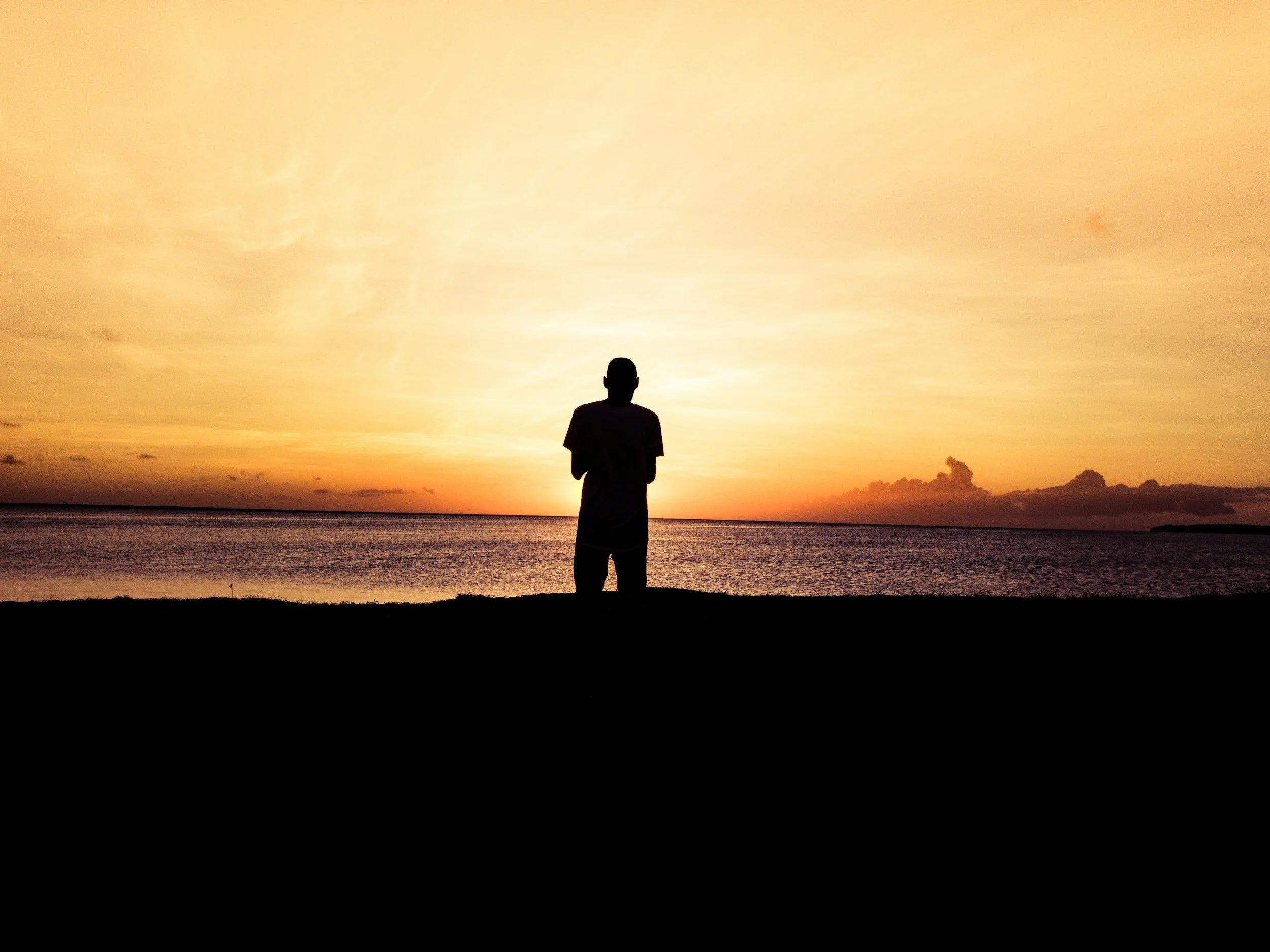 A person standing on the beach during sunset, facing the water and sky with a silhouette against the colorful sky.