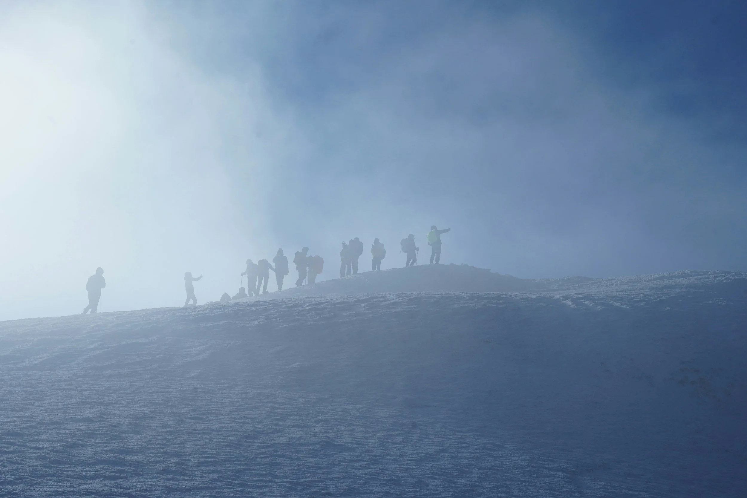 Group of hikers ascending a snowy mountain in foggy weather, some carrying backpacks and a person playing guitar.