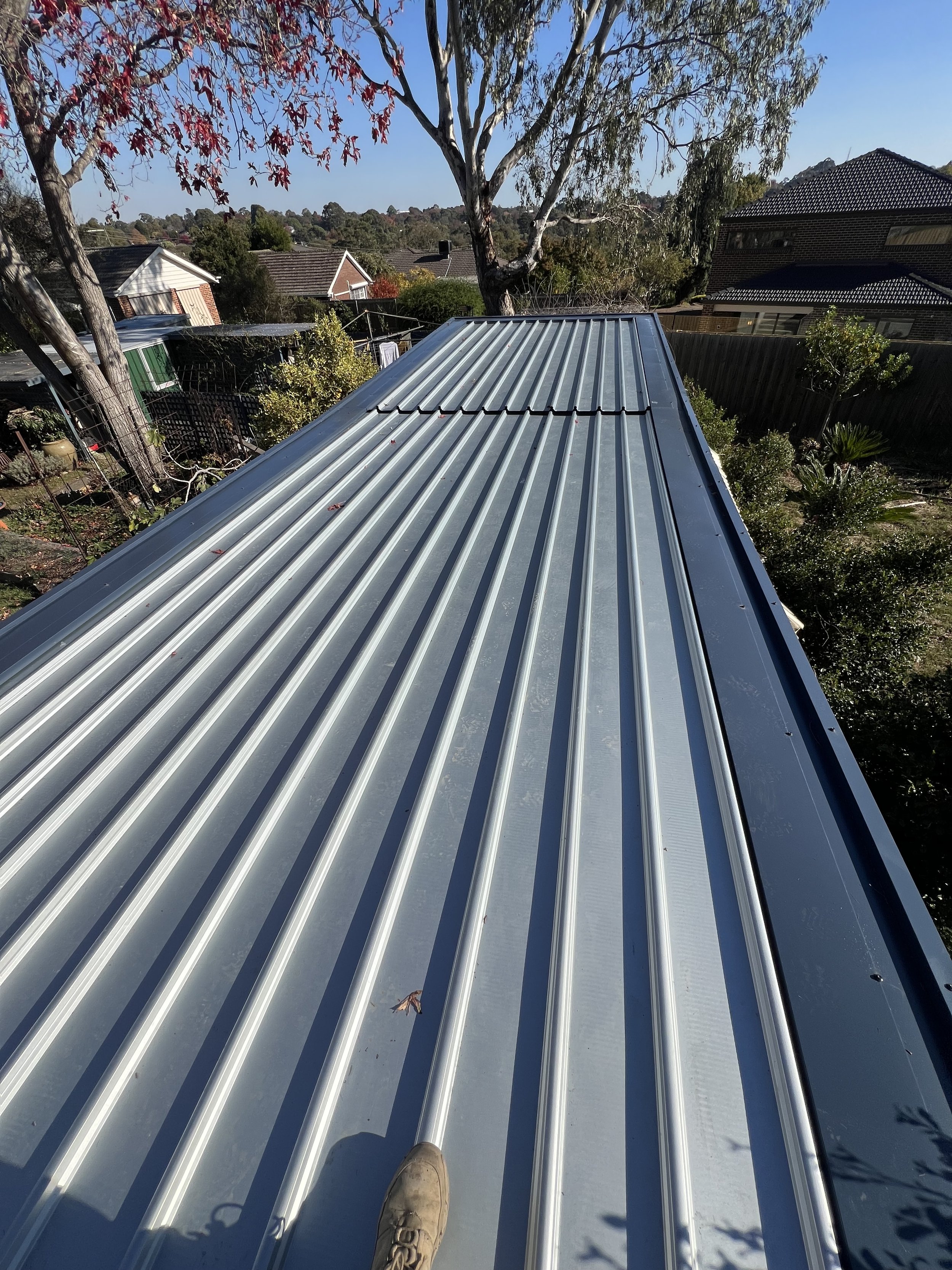 View from a rooftop with metal corrugated roofing, showing a person's feet and shoes at the edge, and a suburban neighborhood with trees, houses, and a clear blue sky in the background.
