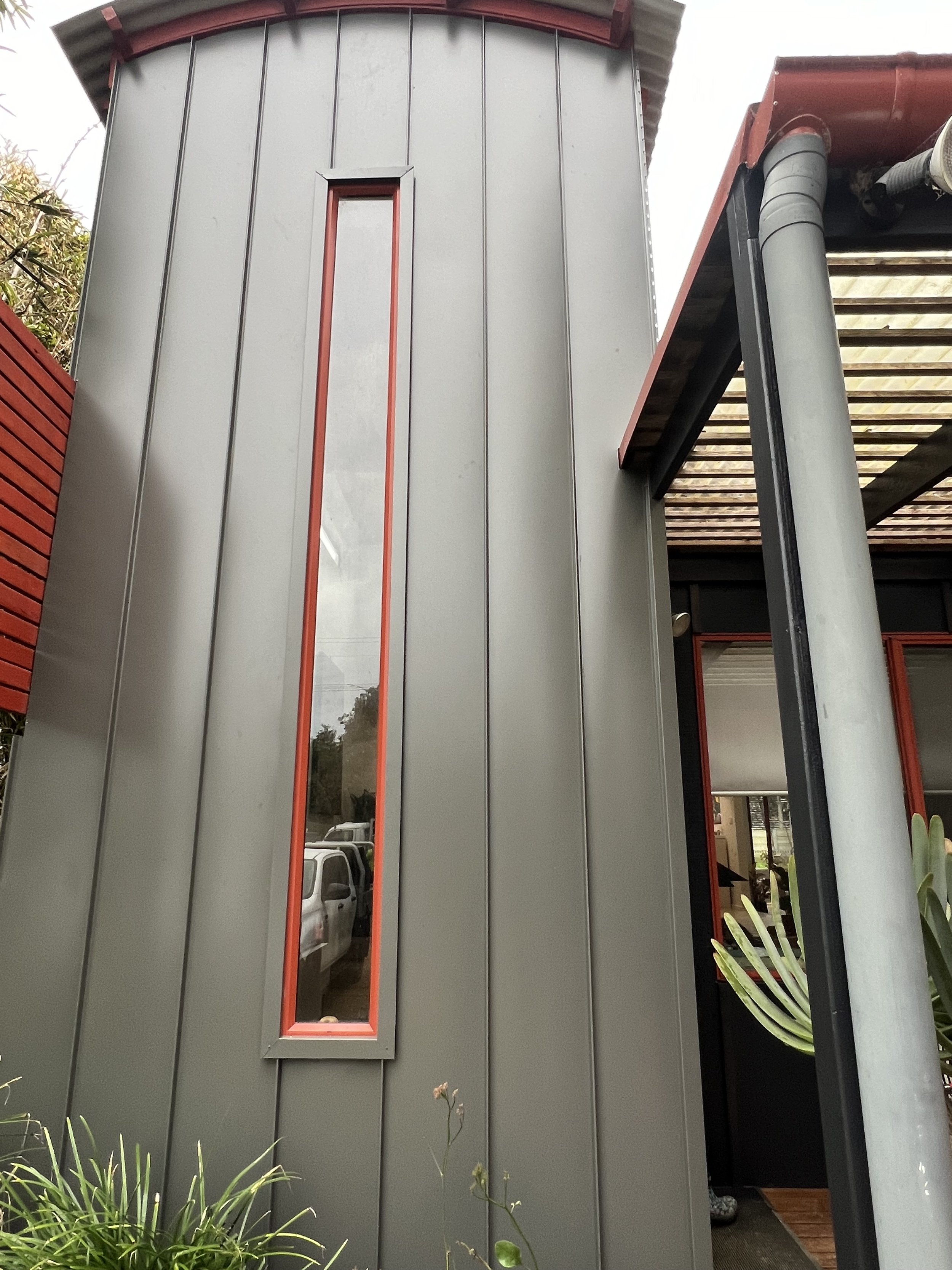 A modern building with gray metal siding, narrow vertical window with red border, and a matching red trim under the roof. A sliding black door is partly open, revealing the interior. There are some green plants and trees nearby.