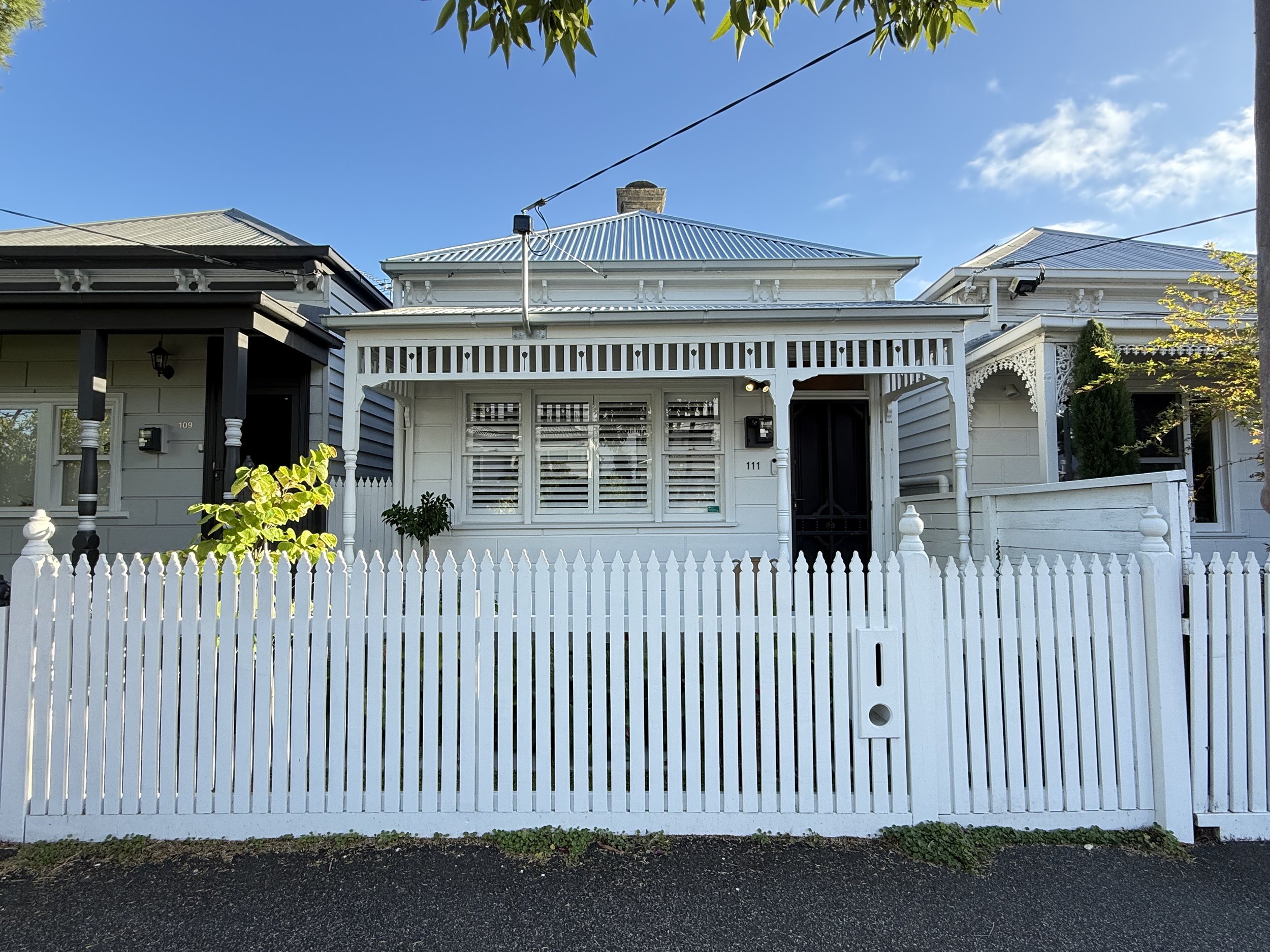 Front view of a white Victorian-style house with a porch, decorative trim, and a porch railing, fenced by a white picket fence, under a blue sky with a few clouds.