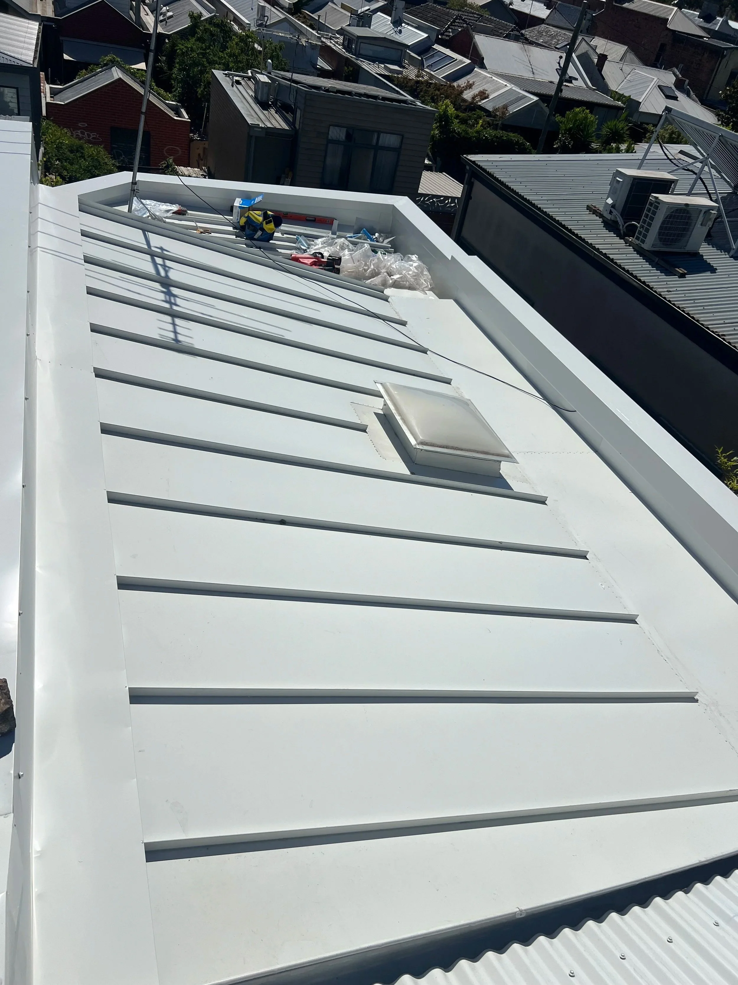 The image shows the roof of a building under construction or repair with construction tools and materials scattered on it, including a level, a power drill, wires, and plastic sheets. Surrounding buildings with various roof styles and HVAC units are 