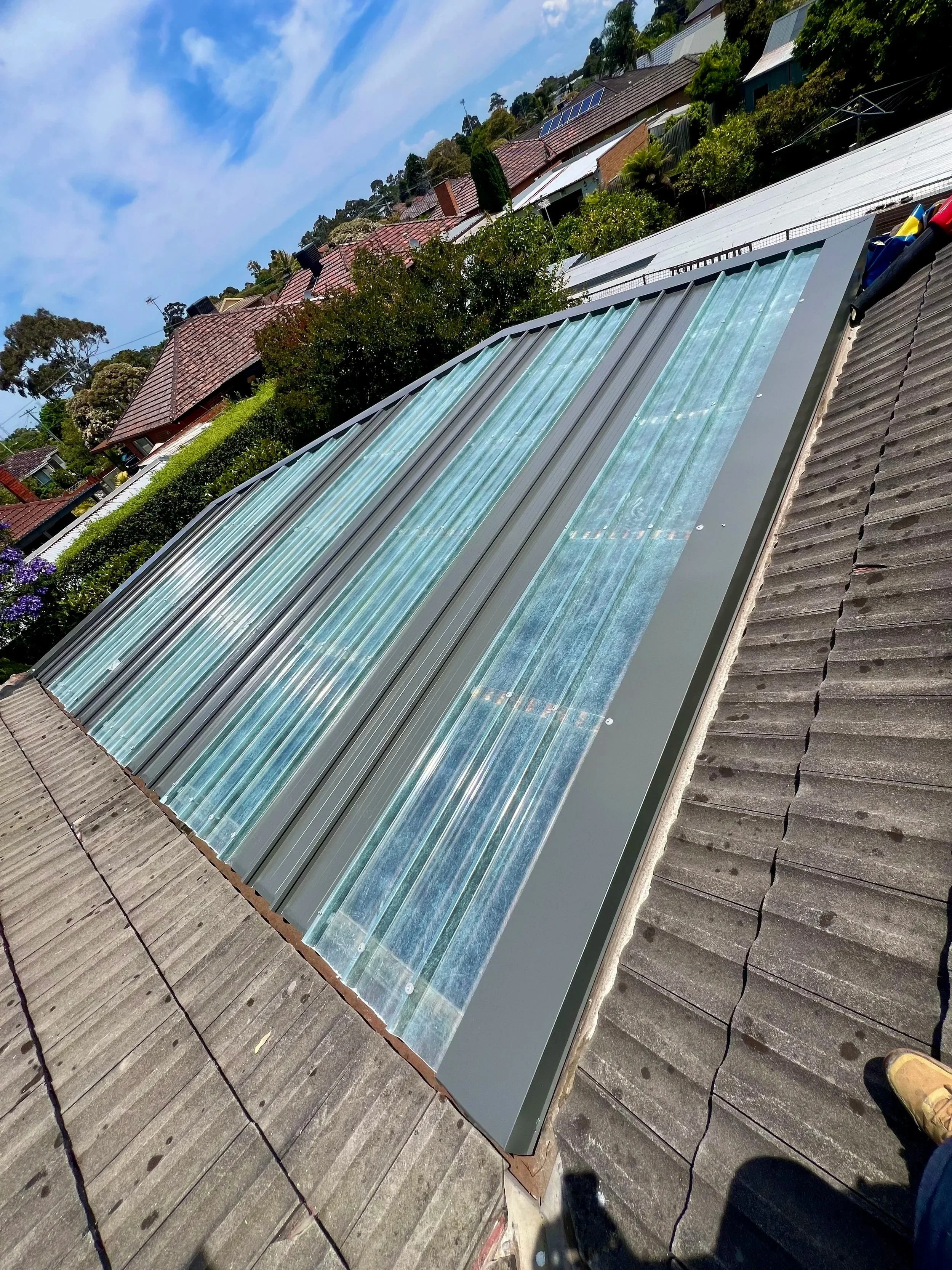 Roof with solar panels and a glass skylight, rooftops and trees in a residential neighborhood under a blue sky with clouds.