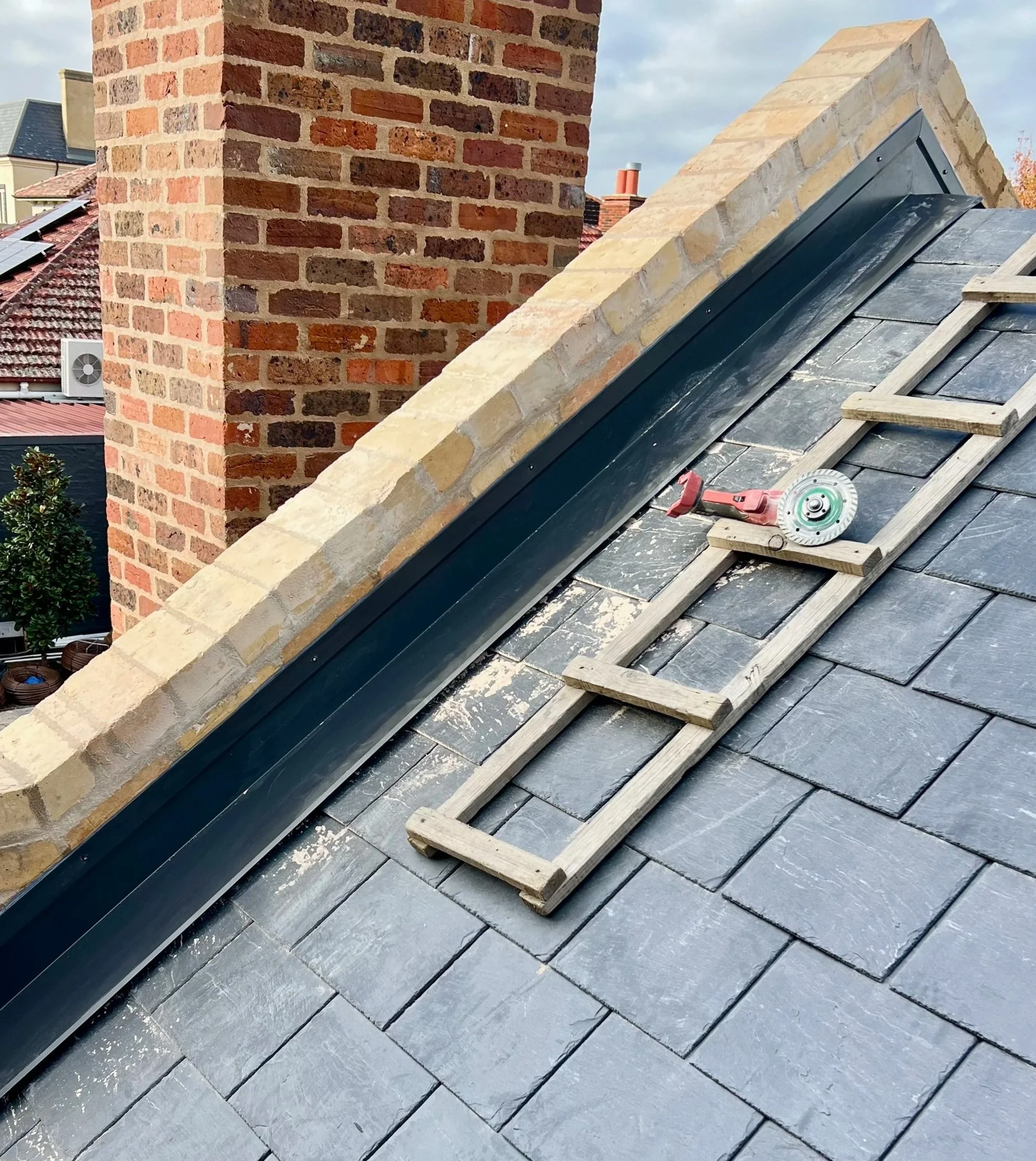 Construction materials and tools on a rooftop, including slate tiles, wooden battens, a circular saw, and a red power drill, with a brick chimney and neighboring houses in the background.
