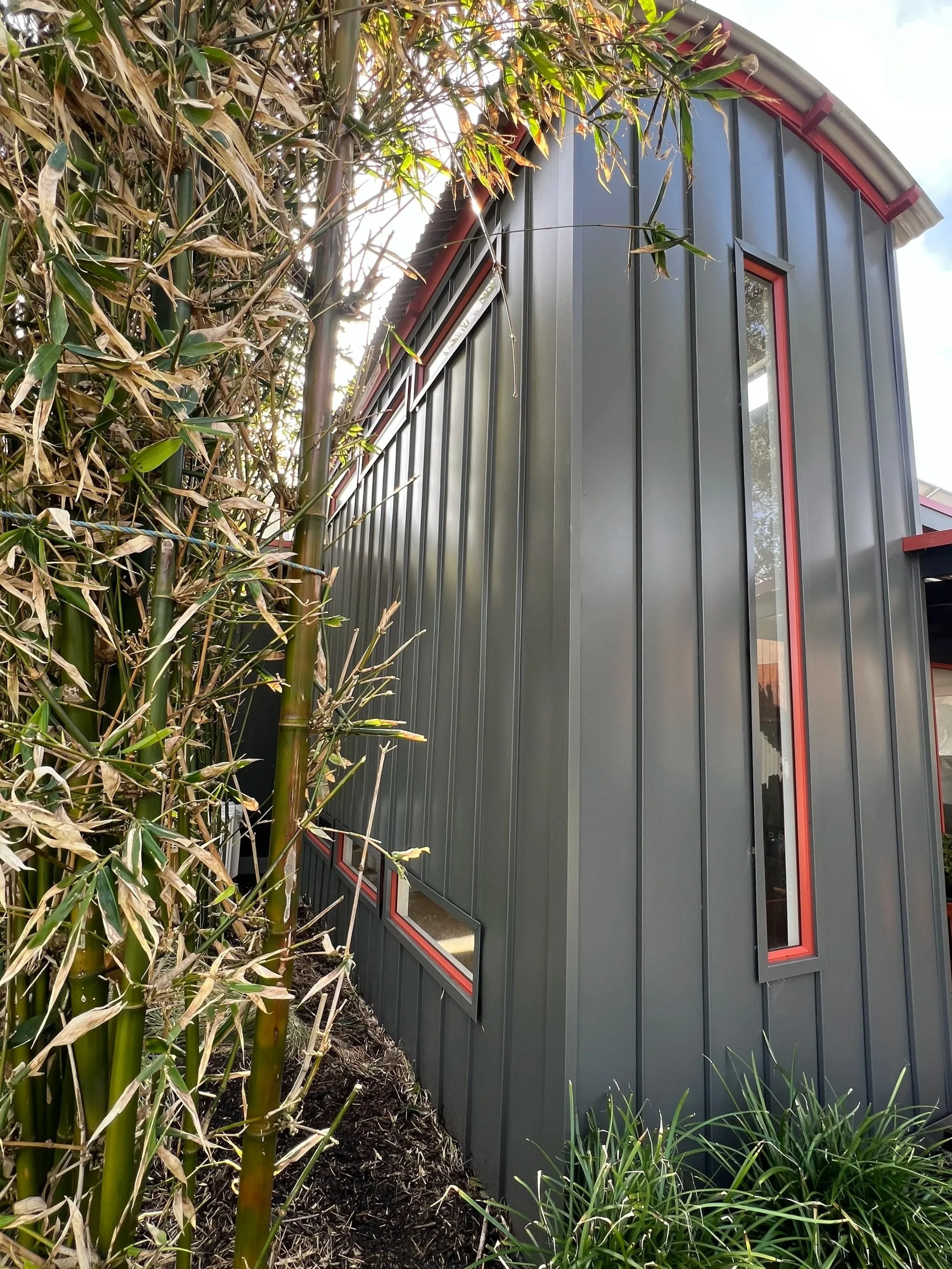 A modern building with vertical metal siding and red-framed windows, situated next to bamboo plants and other green vegetation.