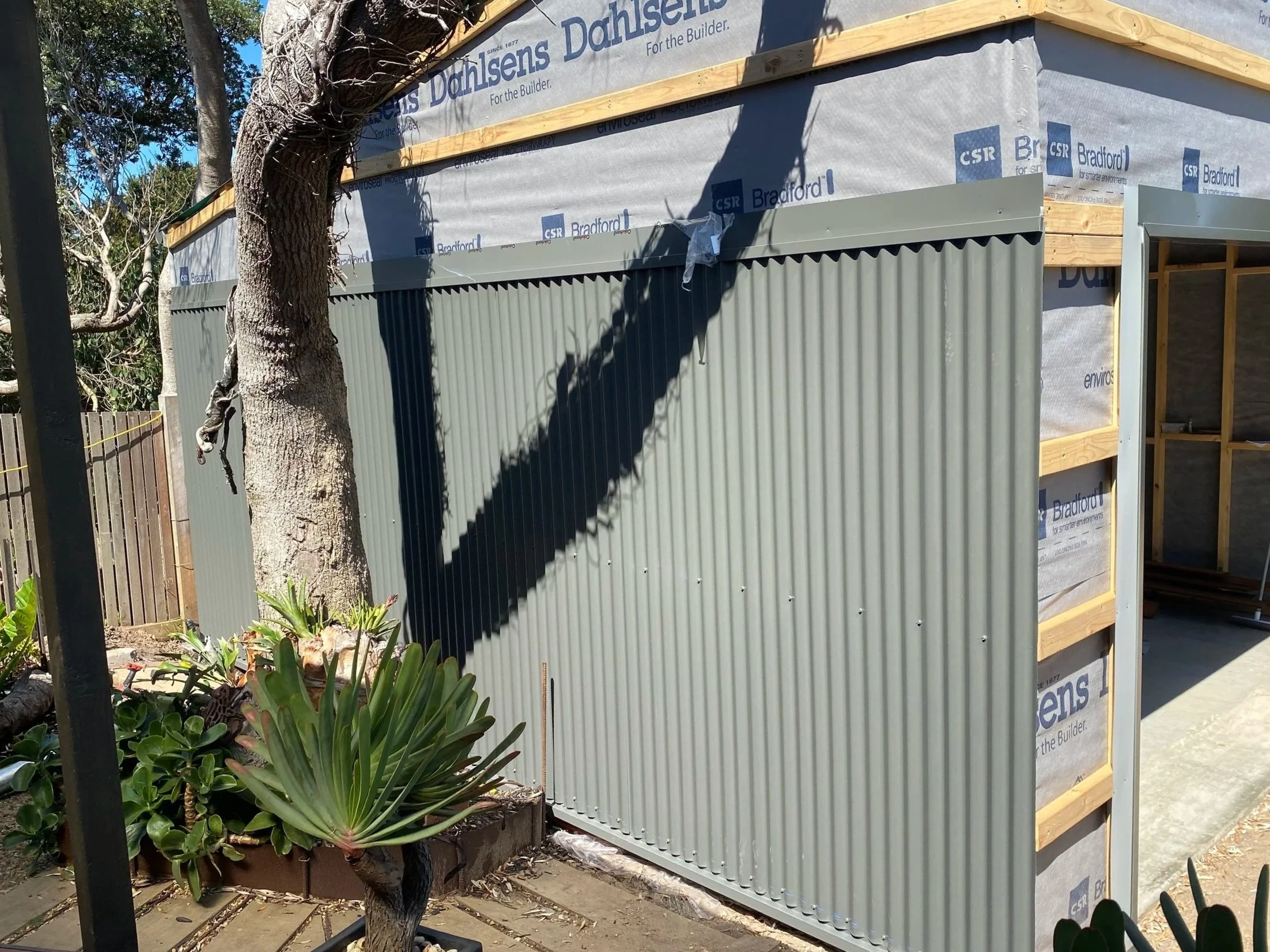Construction site with a partially built structure, a tree in the foreground, and a wooden fence. Building wraps with 'Bradford' and 'CSR' logos are visible.
