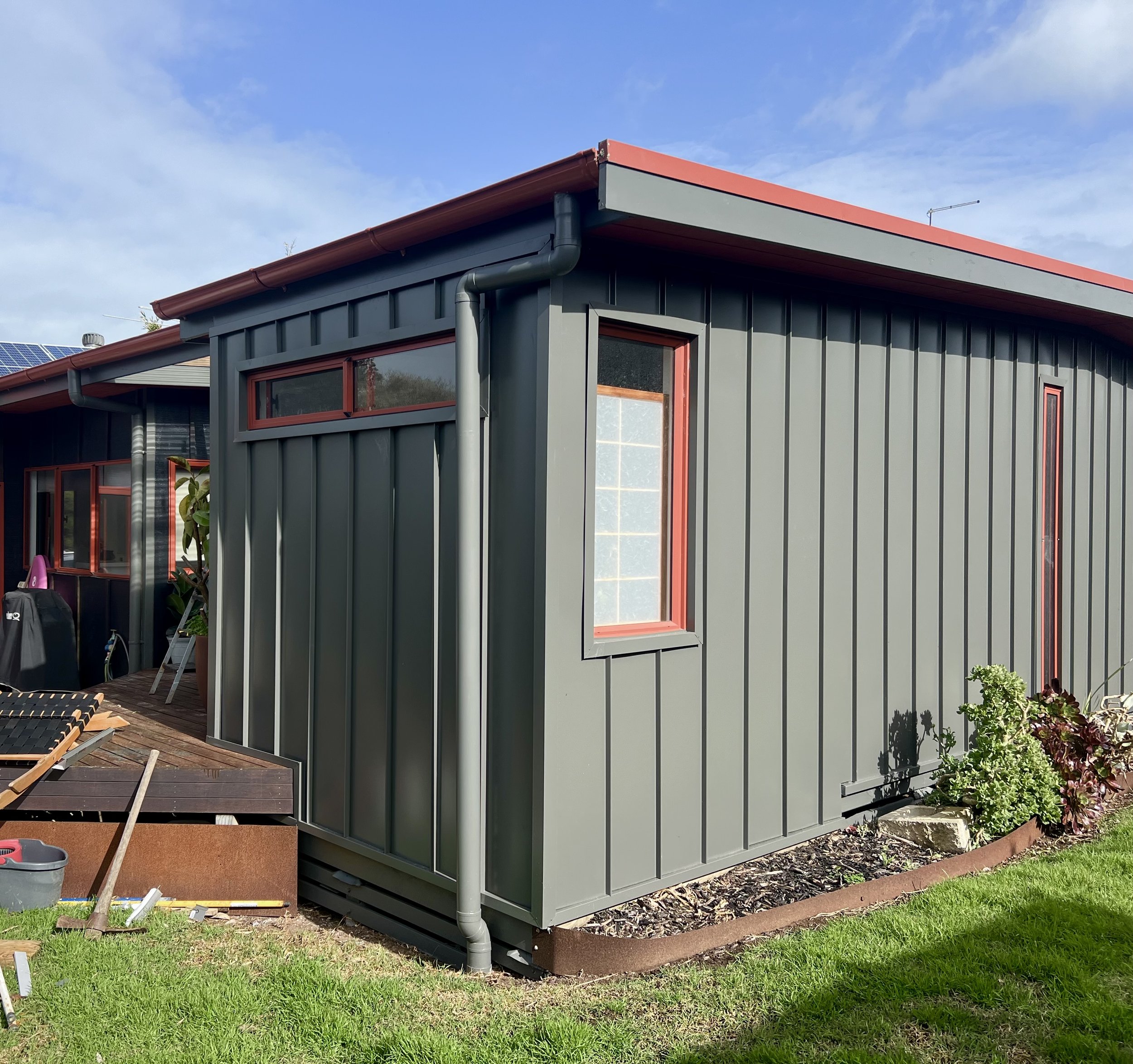 A modern gray house with red window and roof accents, seen from the side under a blue sky with some clouds. There's a garden with green grass and some plants nearby.