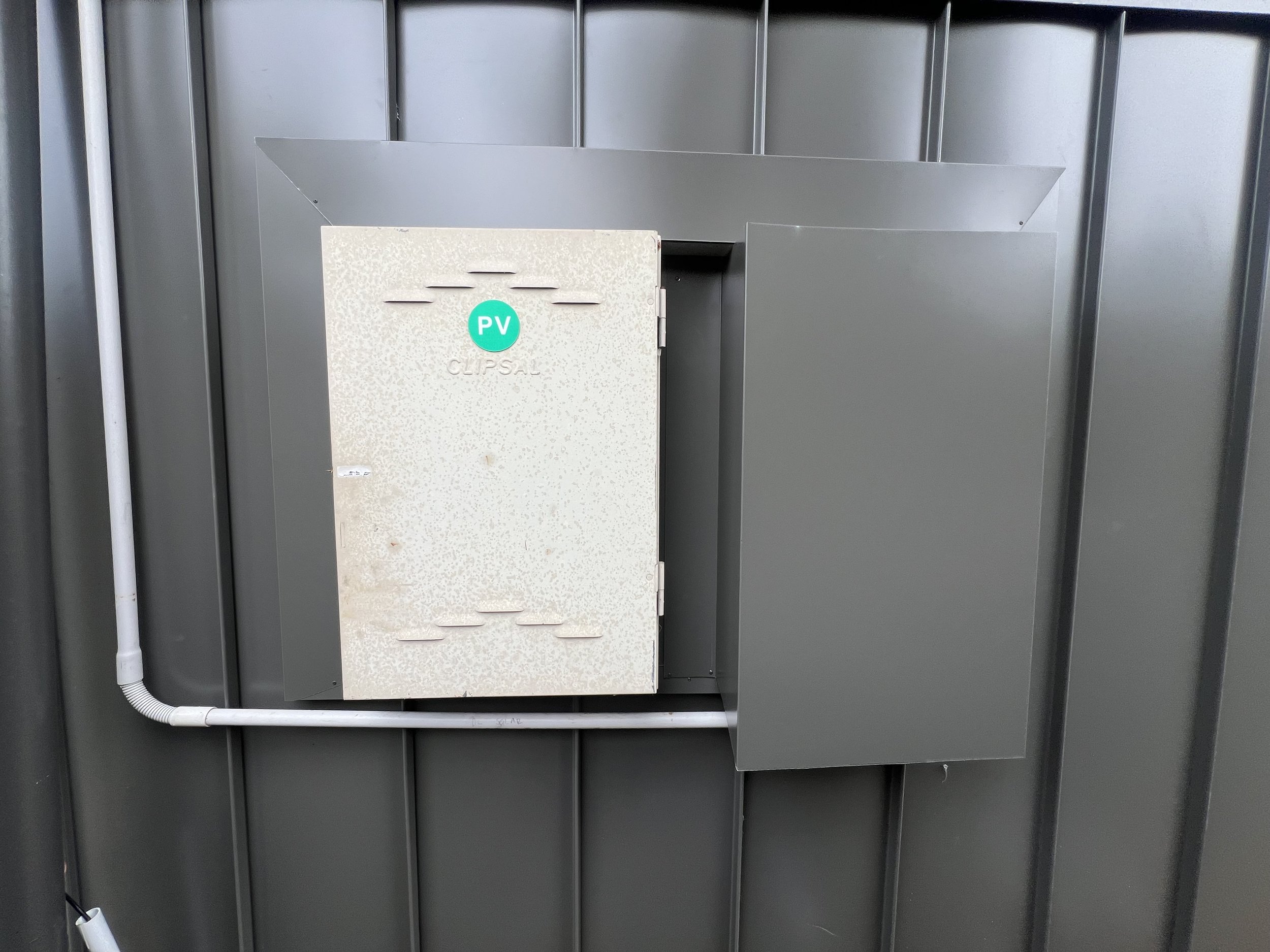 Electrical equipment box with PVC label on it, mounted on a metallic wall panel with visible piping.
