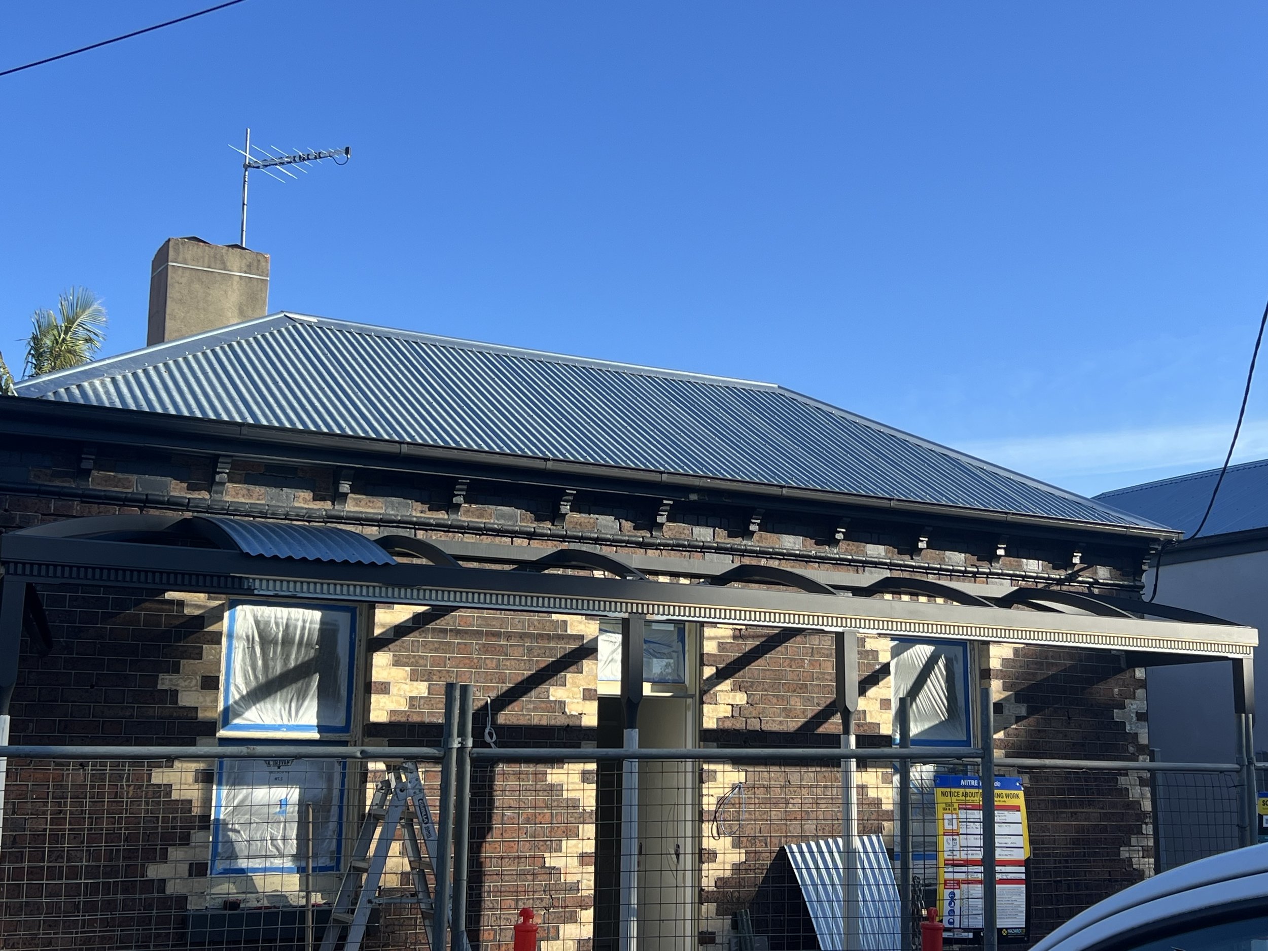 Construction site with a house under renovation, showing a brick wall, a corrugated metal roof, windows covered in plastic, scaffolding, and safety signs.