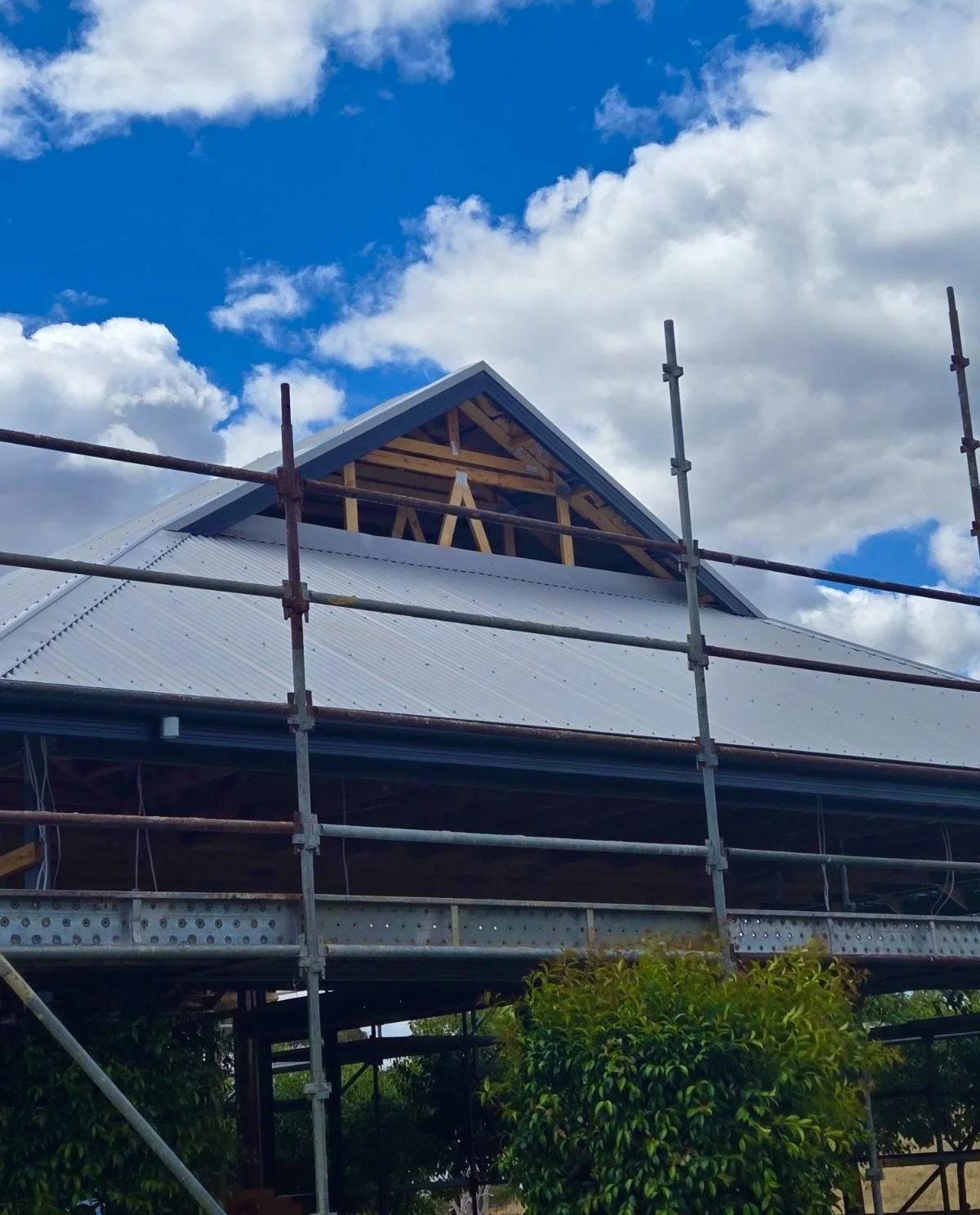 Construction site with scaffolding around a house, roof partially completed with visible wooden framing, under a blue sky with clouds.