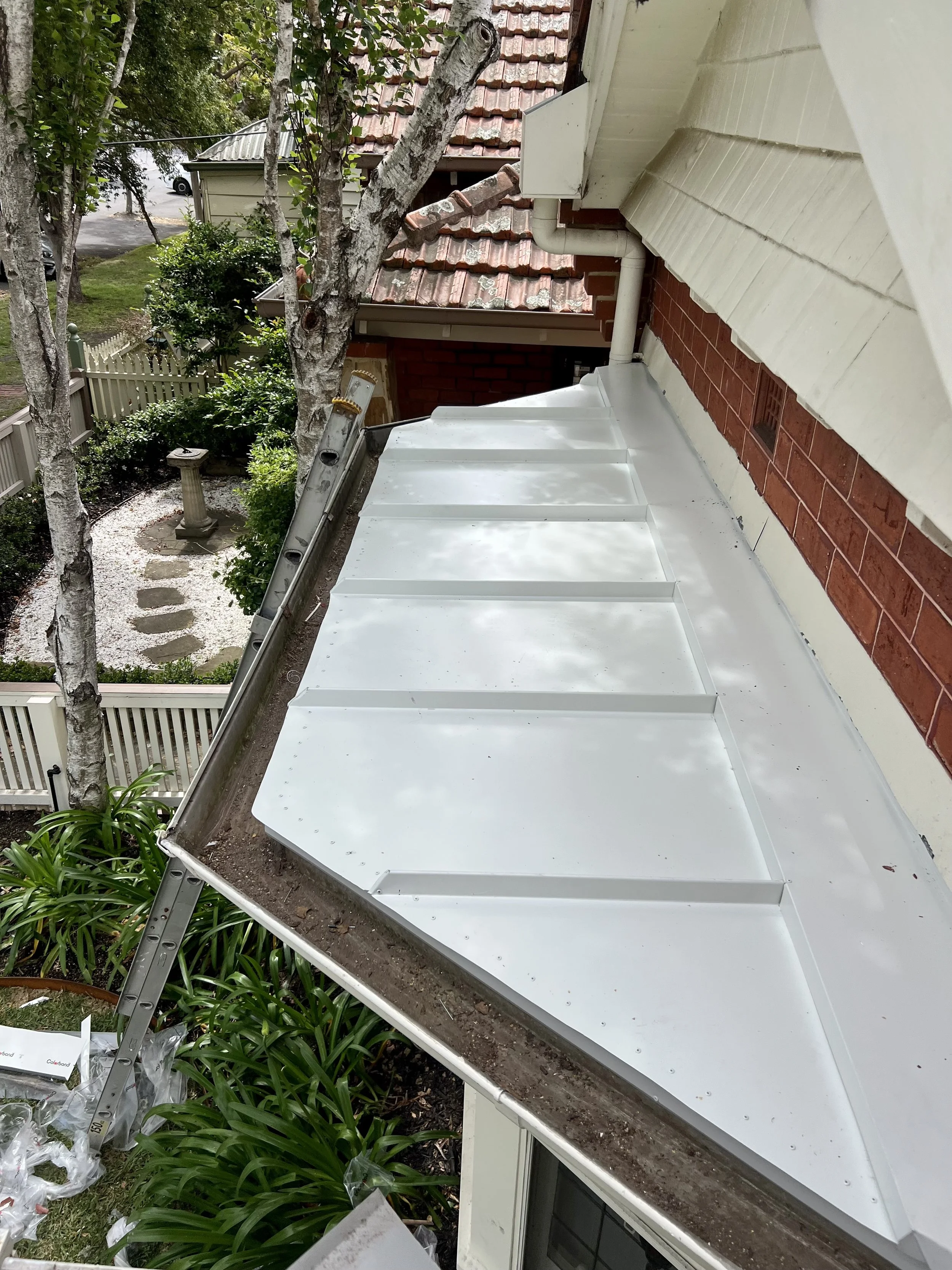 View of a house roof with a white metal awning, a tree, and garden plants in the yard.