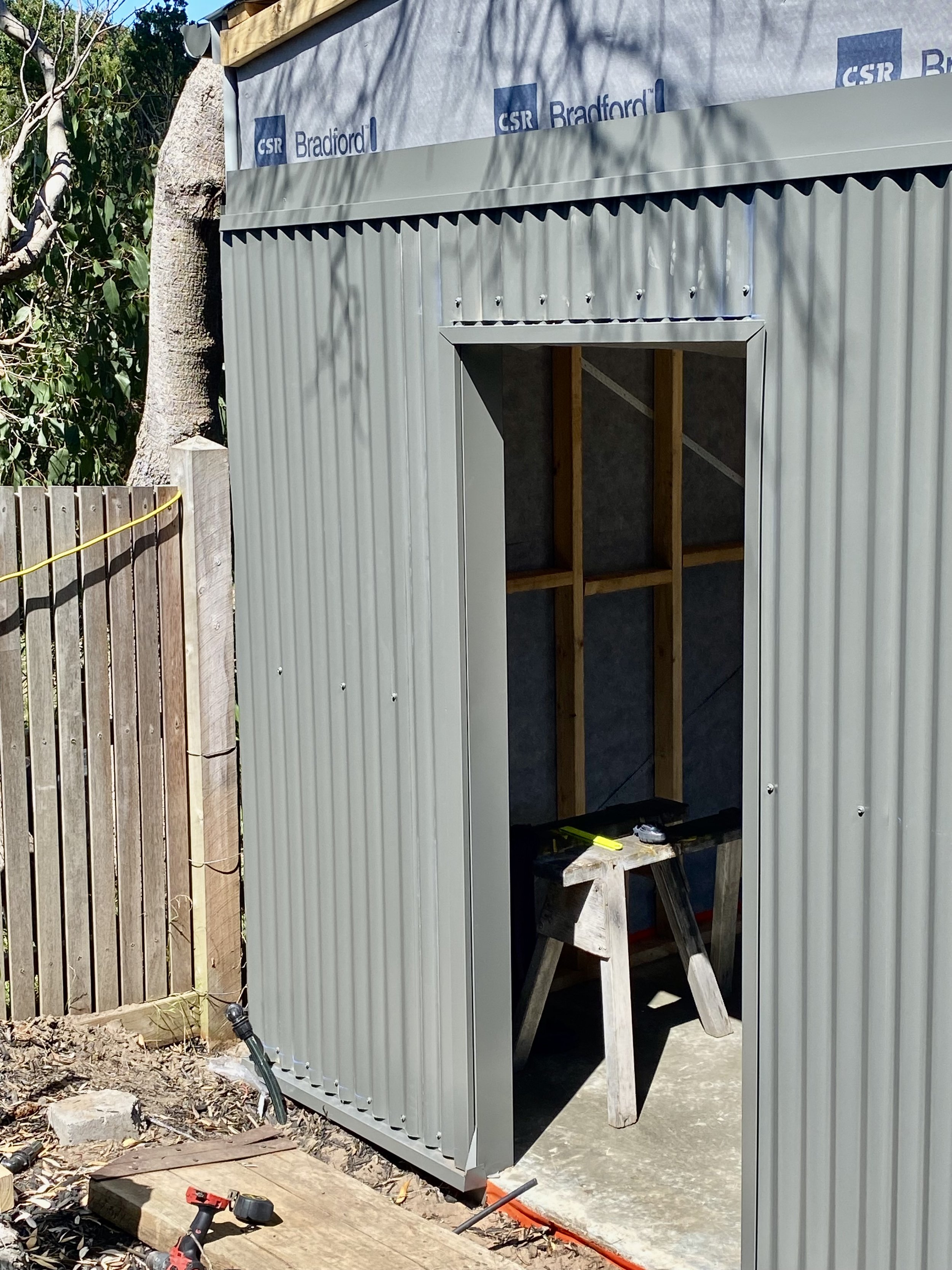 Construction area with a partially built metal shed structure, wooden support beams, and construction tools on a workbench. There is a round tree and a wooden fence nearby.