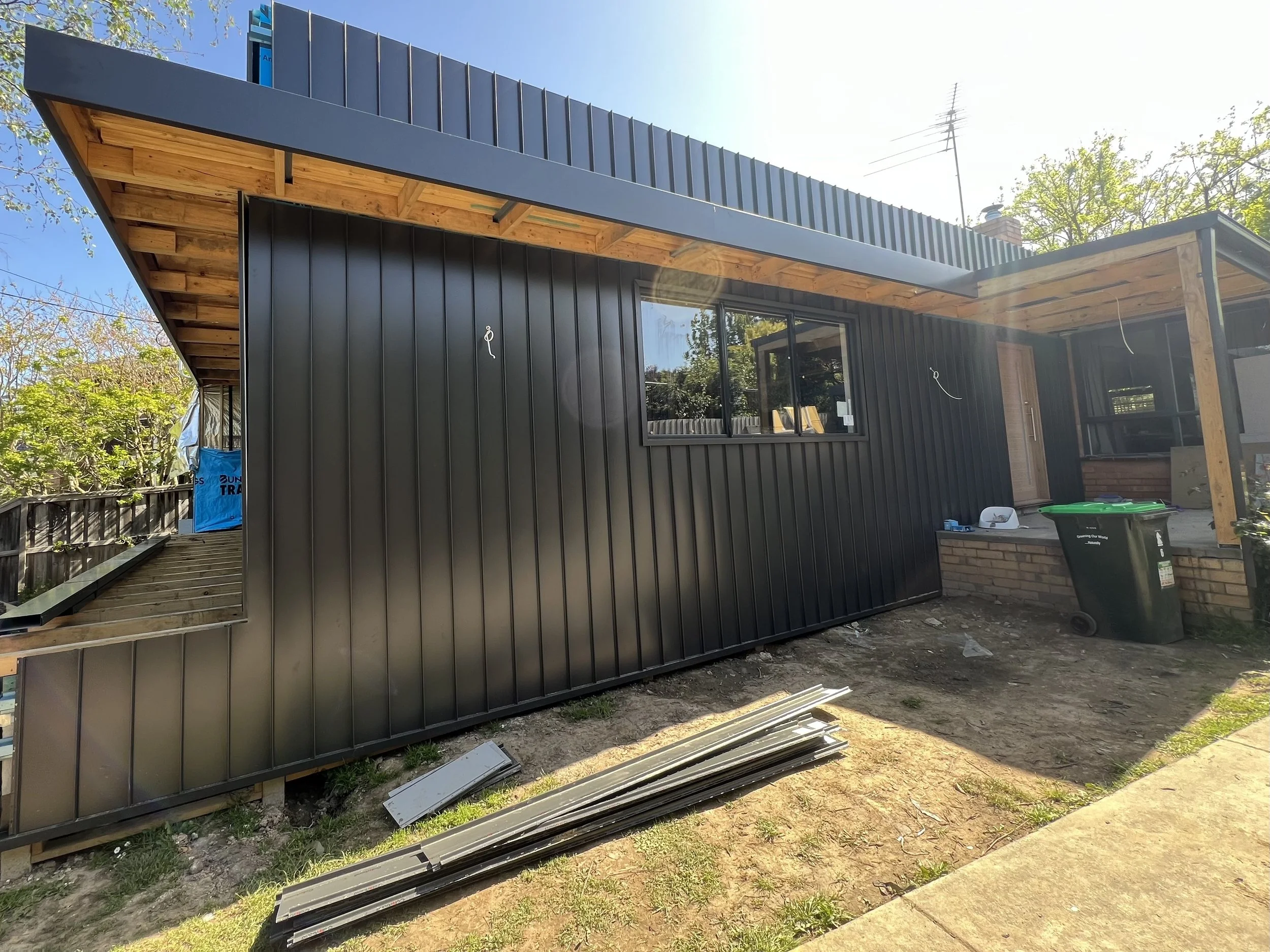 Exterior view of a house under construction with black metal siding, wooden fascia, a window, and construction materials on the ground.