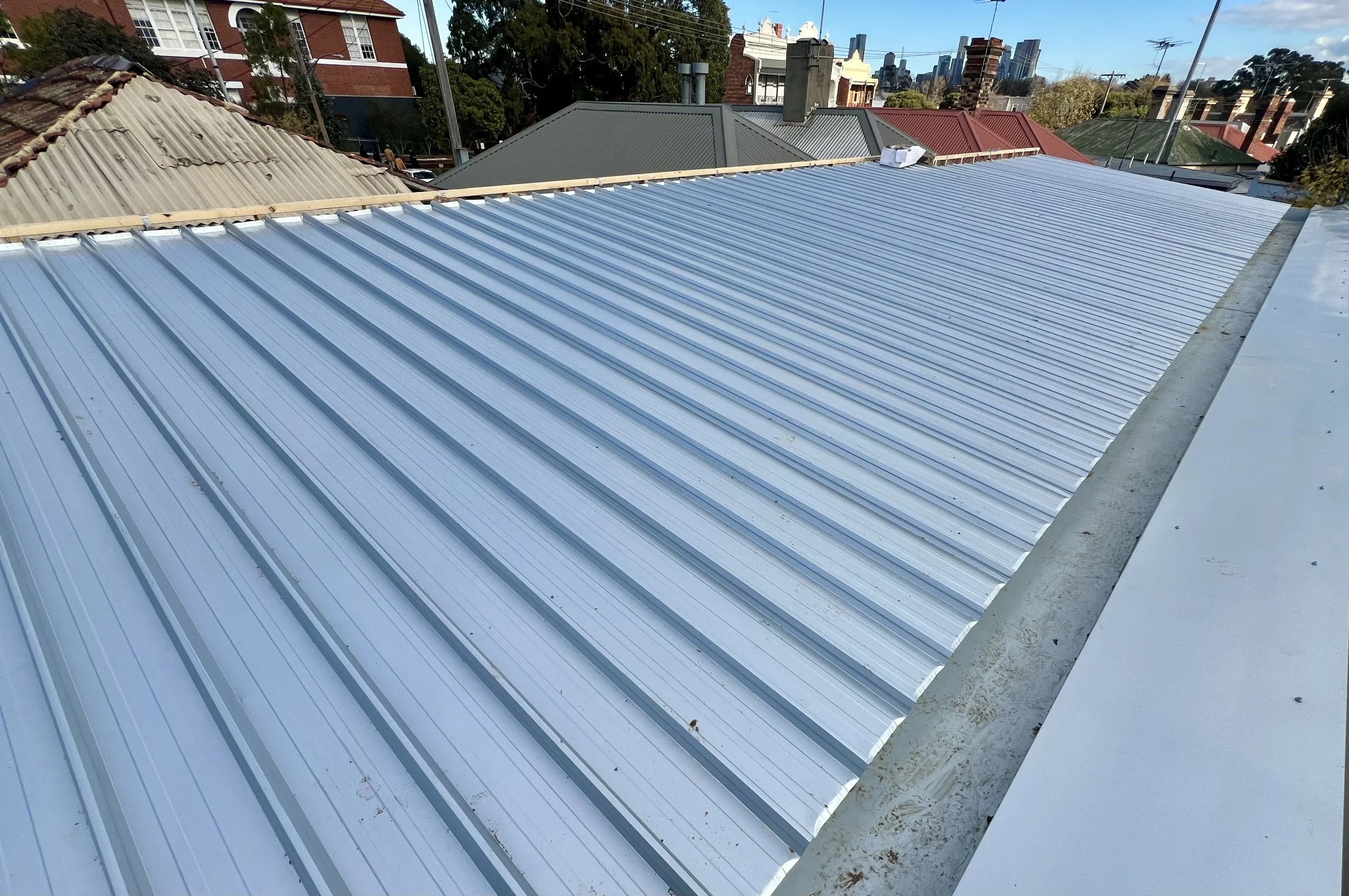 A rooftop with new silver metal roofing panels installed, adjacent to older roofs with tile and metal materials, in an urban neighborhood with buildings and trees in the background.