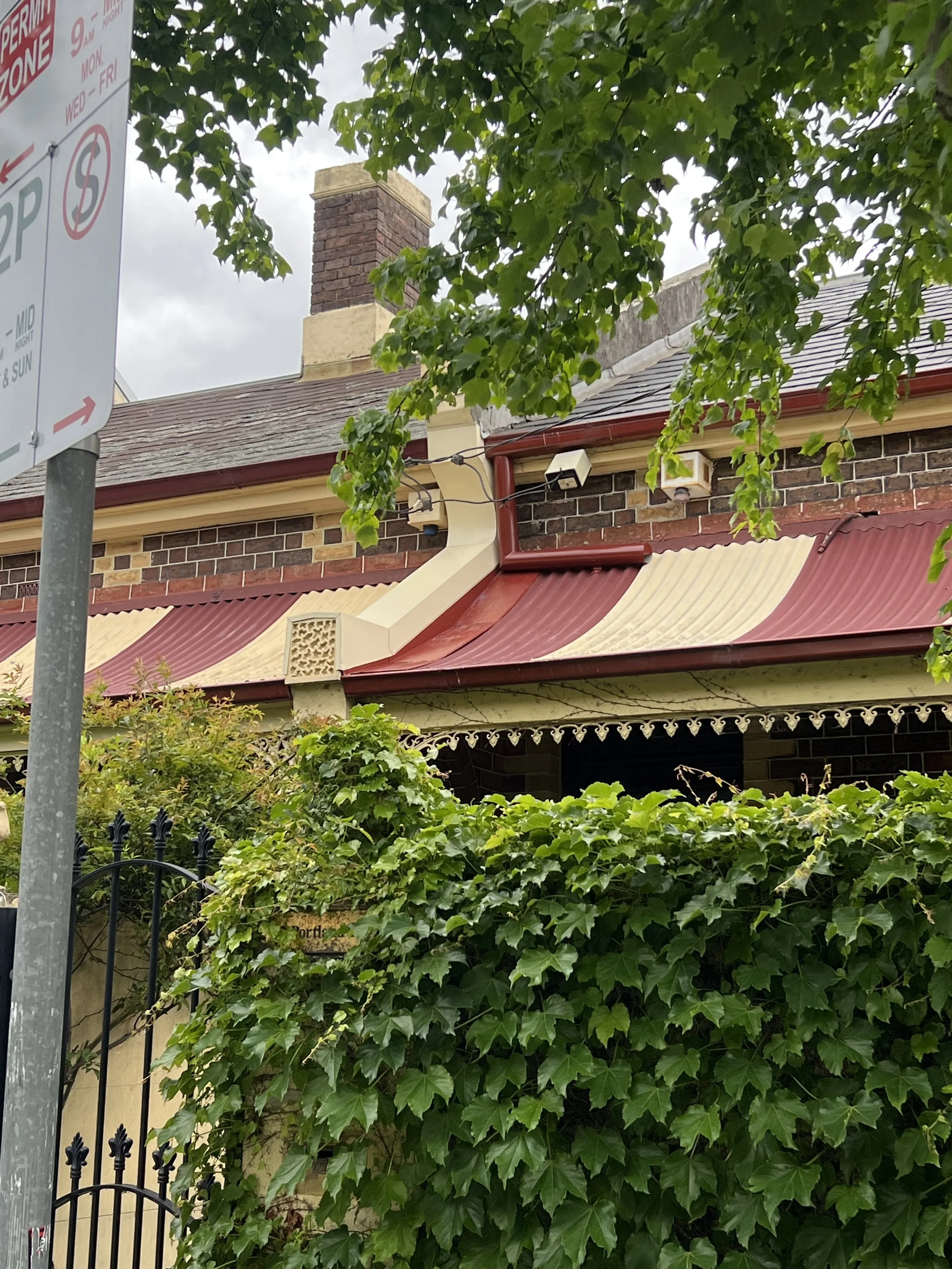 The photo shows a red brick house with a red and cream roof, a chimney, and a wall covered with lush green ivy. A street sign is partially visible on the left side of the image.