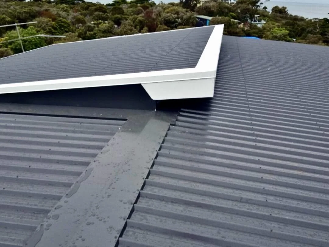Close-up view of a metal roof with a white solar panel installed on top, surrounded by trees and greenery.