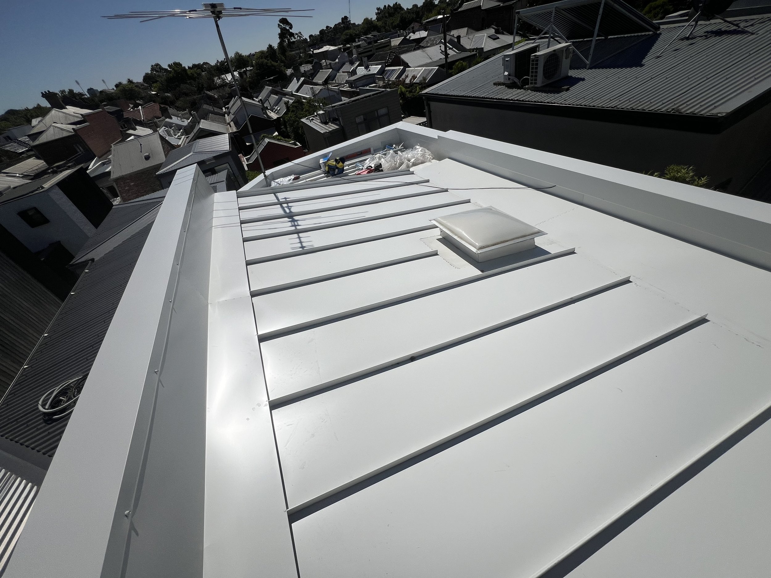 Looking down on a white flat rooftop with a skylight, rooftop equipment, and some construction materials, with neighboring rooftops and trees in the background under a clear sky.